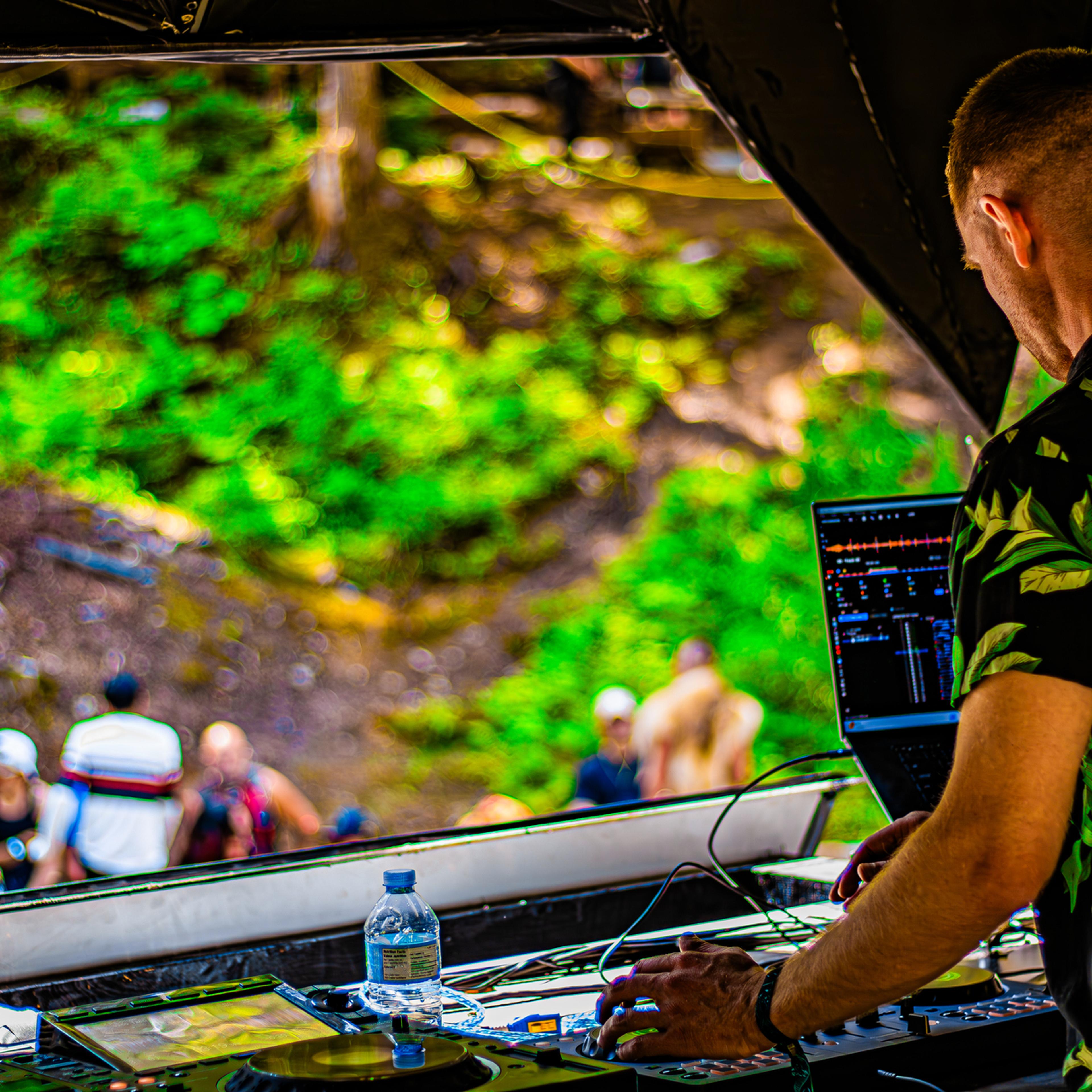 A DJ in a floral shirt works the decks outdoors, lush green forest and seated crowd blurred behind him.