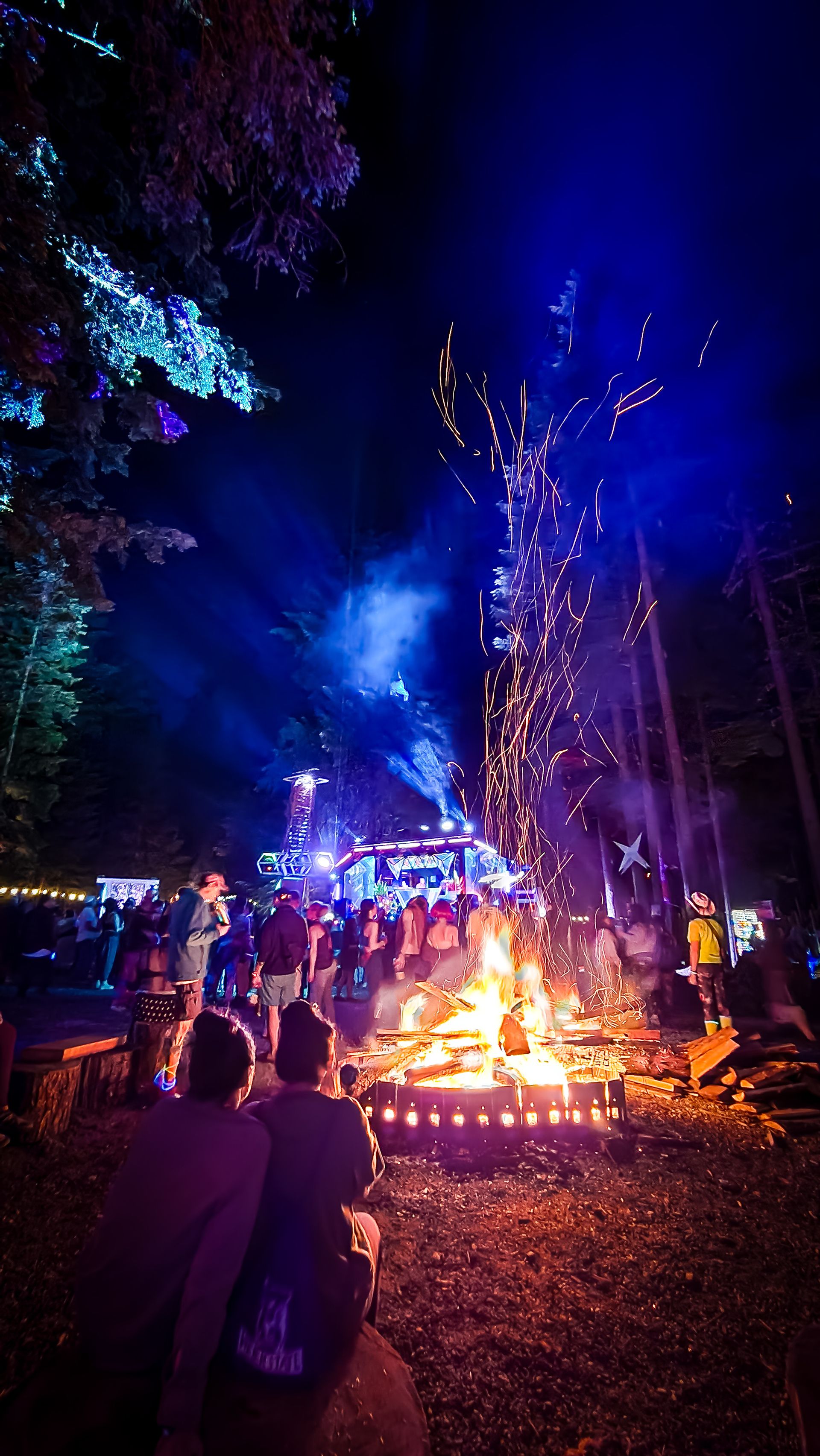 Festival crowd gathered around a roaring bonfire at night, sparks flying, with a glowing blue stage and lit trees in the background.