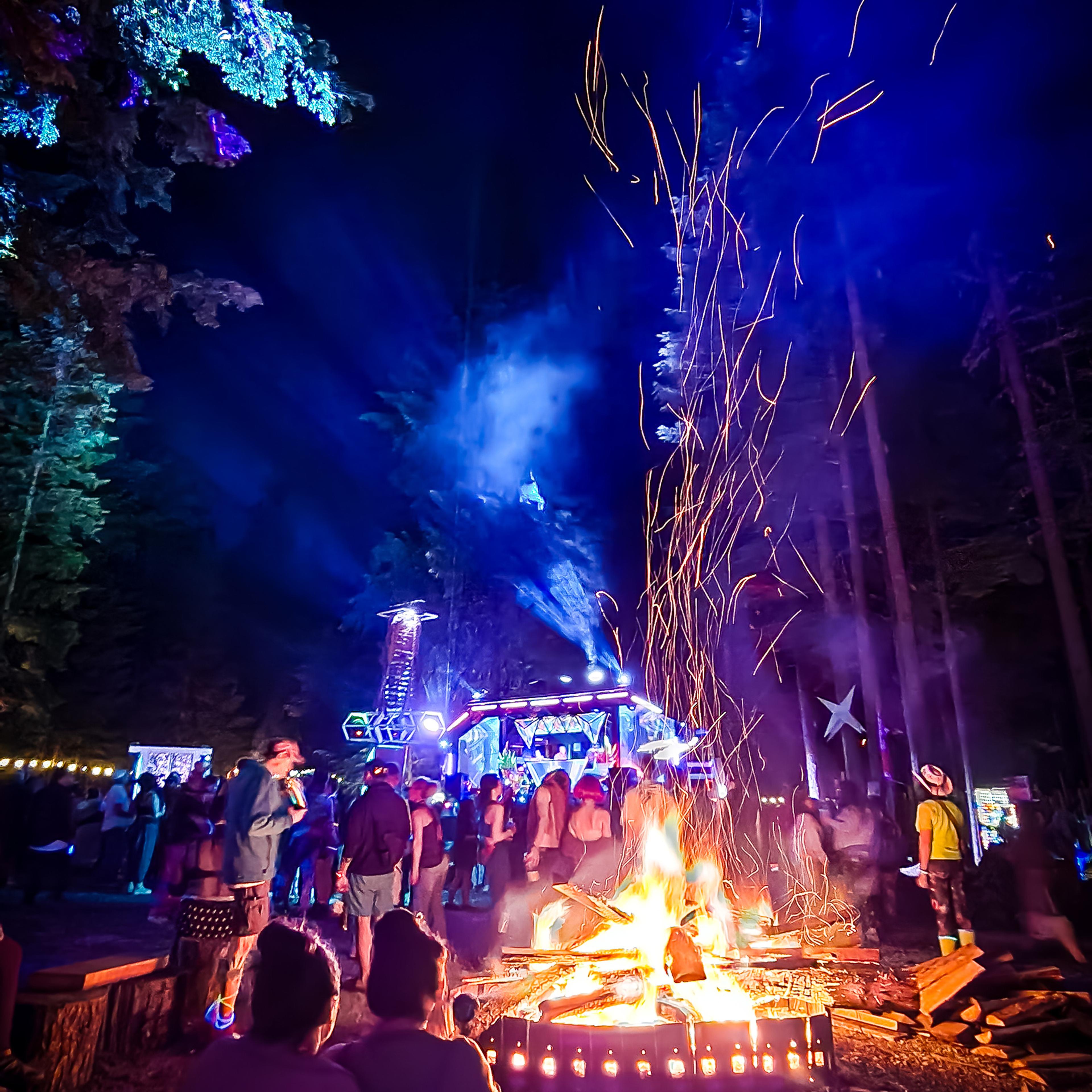 Festival crowd gathered around a roaring bonfire at night, sparks flying, with a glowing blue stage and lit trees in the background.