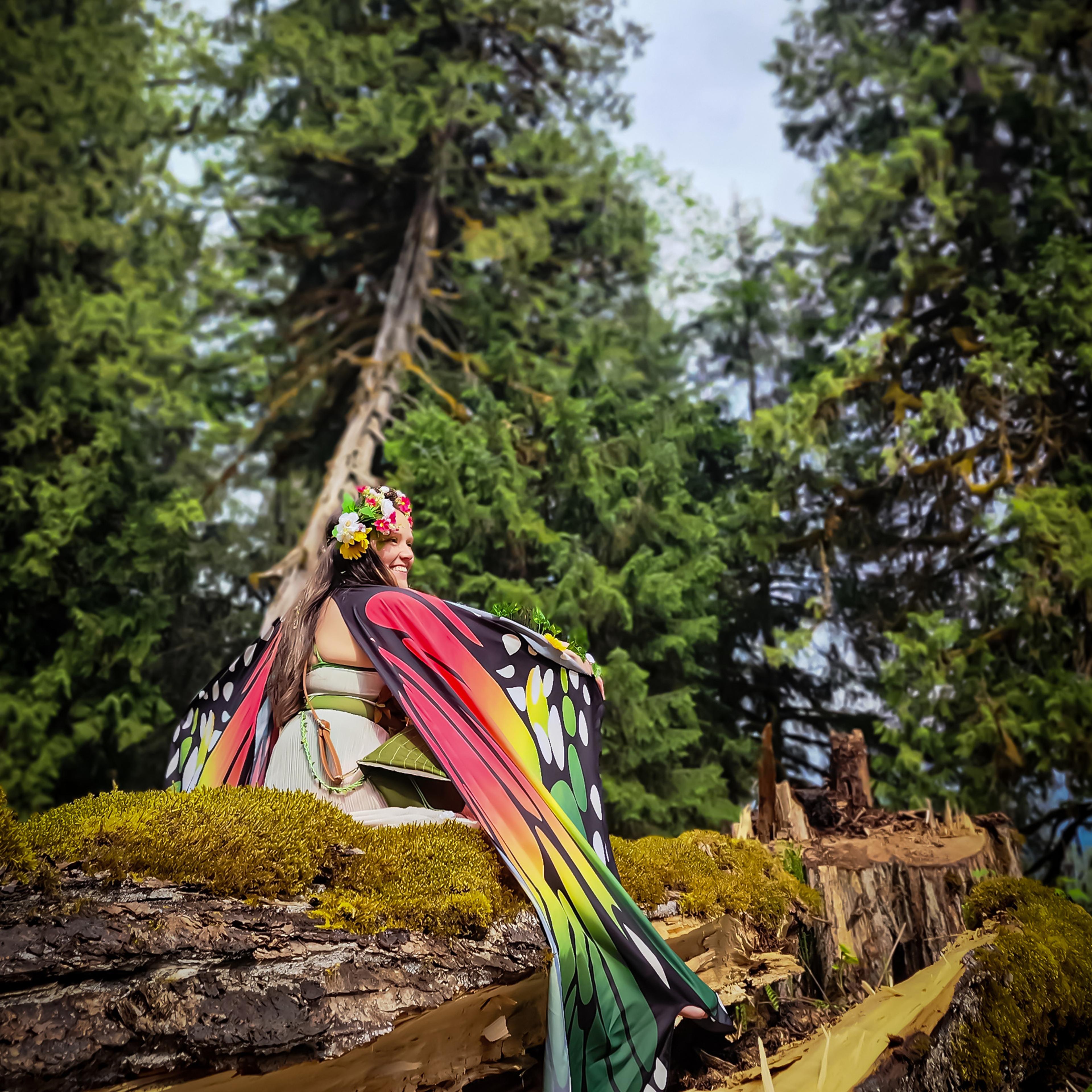 A person in butterfly wings sitting on a large fallen mossy log looking toward tall trees.