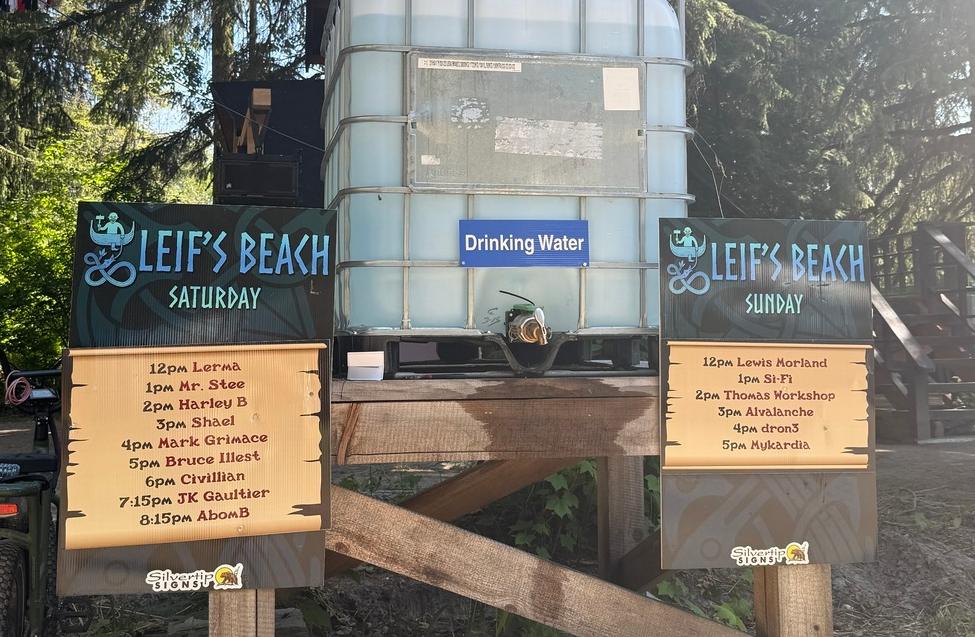 A large drinking water tank stationed between two wooden stage schedule signs for "Leif's Beach" Saturday and Sunday lineups, surrounded by tall evergreen trees.