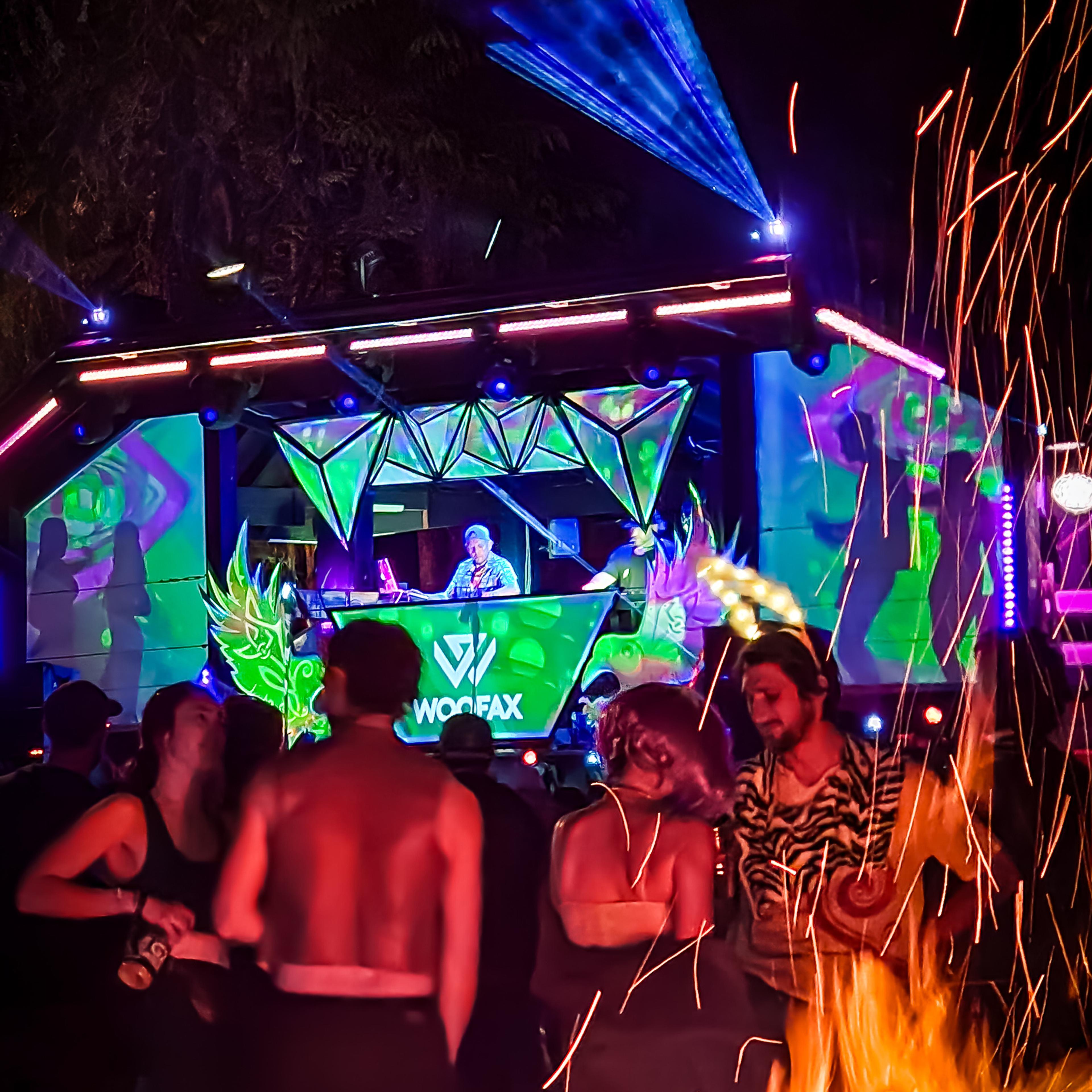 DJ Woofax performing on a vivid green and purple geometric stage with laser lights, bonfire sparks flying in the foreground.