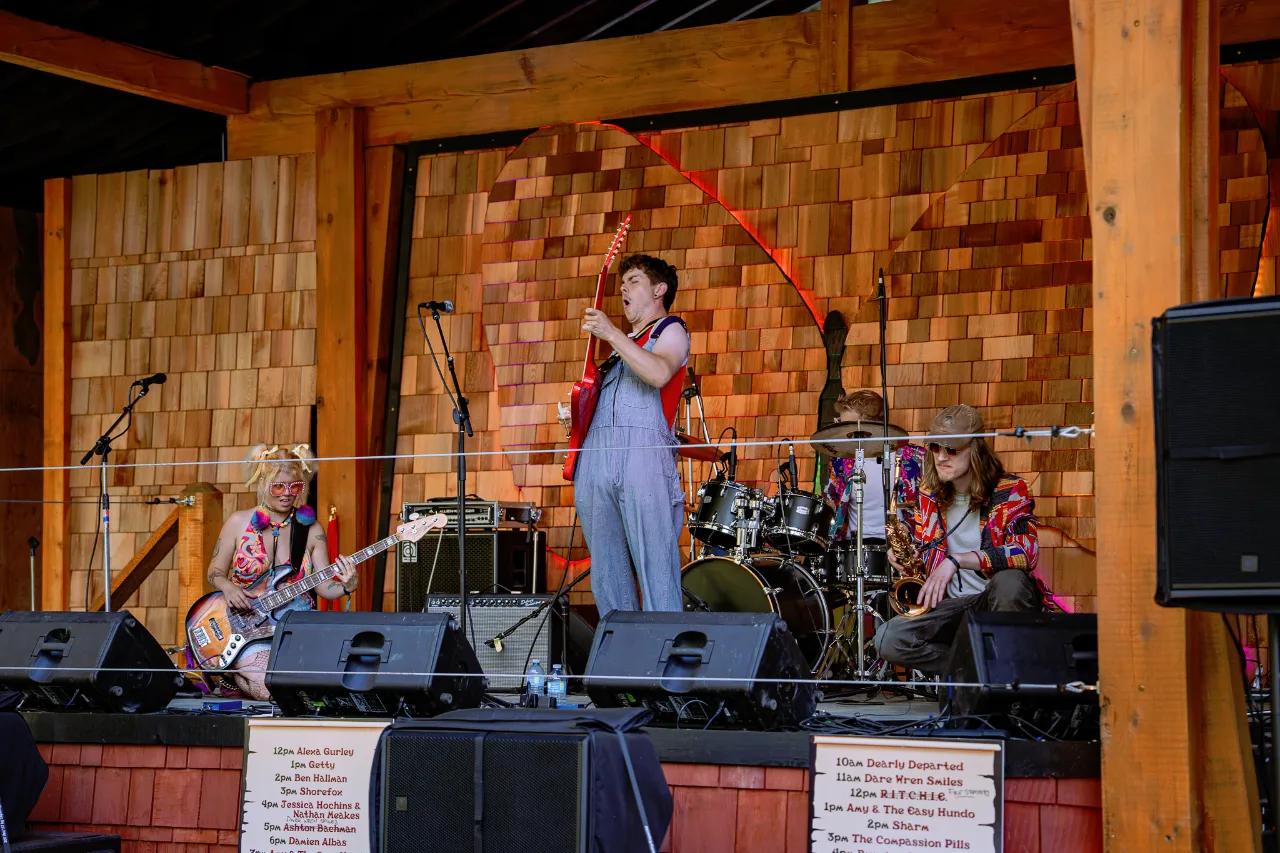 Band performing on AgeHa stage with cedar shingle backdrop and red guitar.
