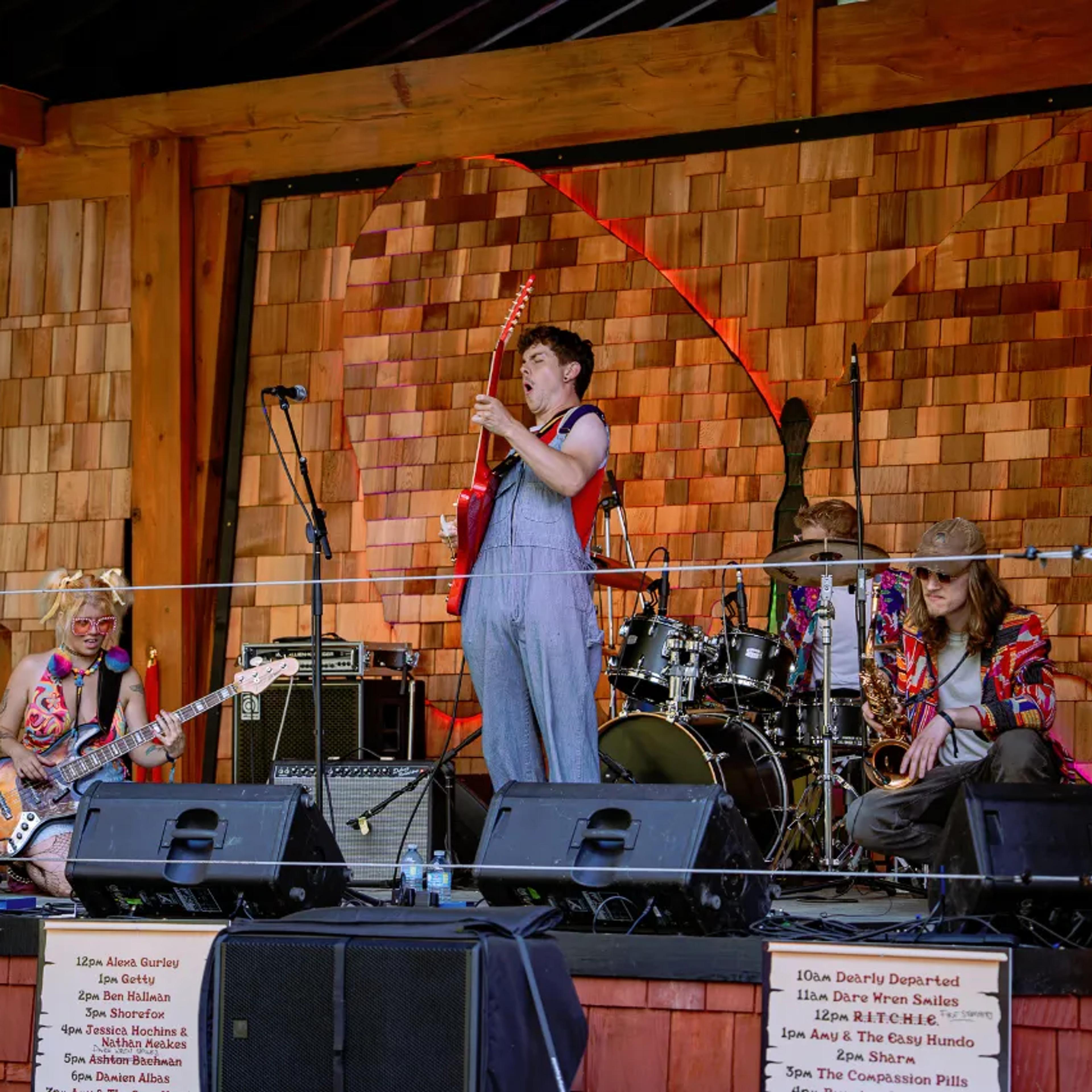 Band performing on AgeHa stage with cedar shingle backdrop and red guitar.