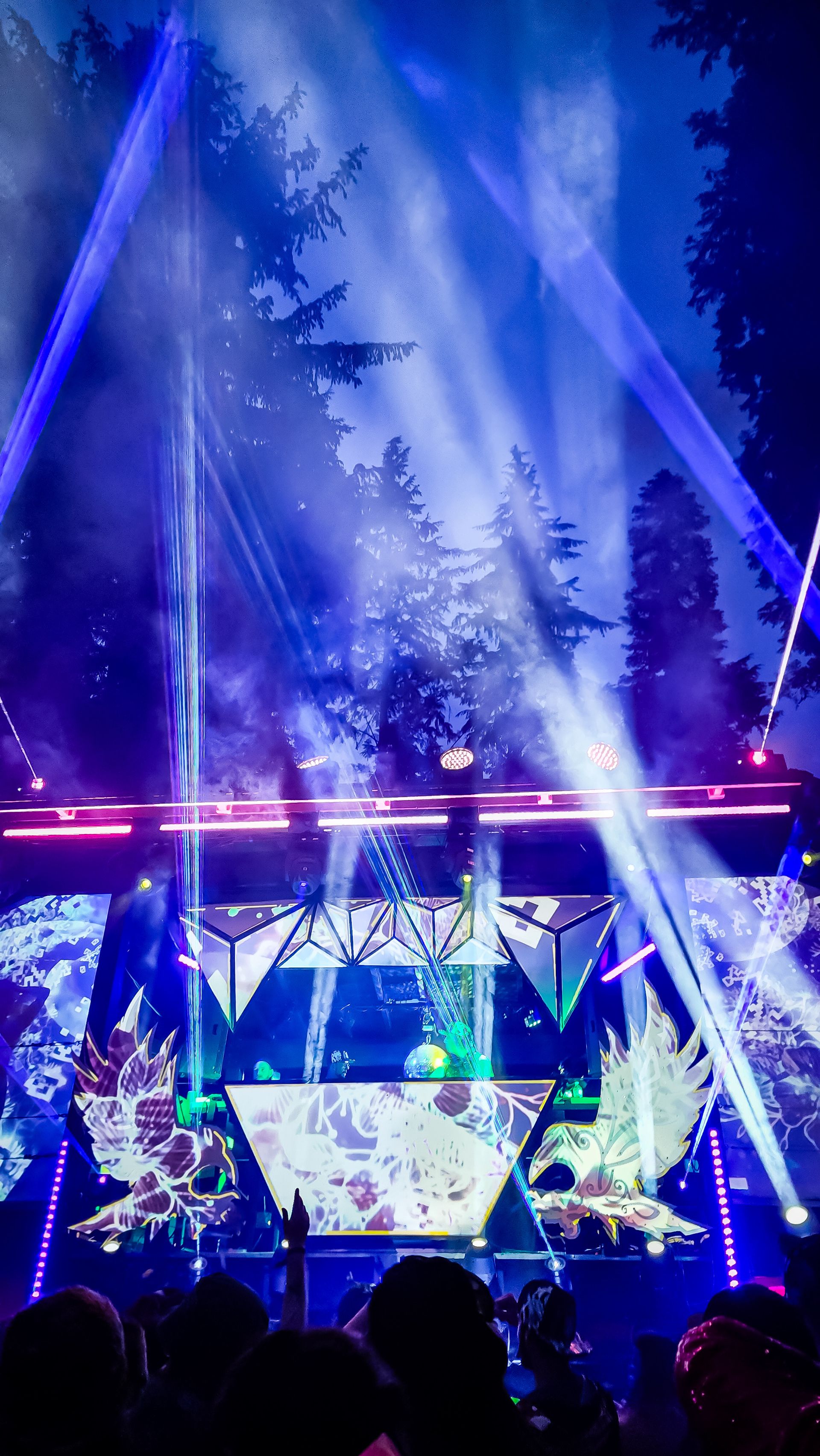 A geometric festival stage bathed in blue and purple laser beams cutting through fog above a silhouetted crowd, with tall trees lit up in the background.