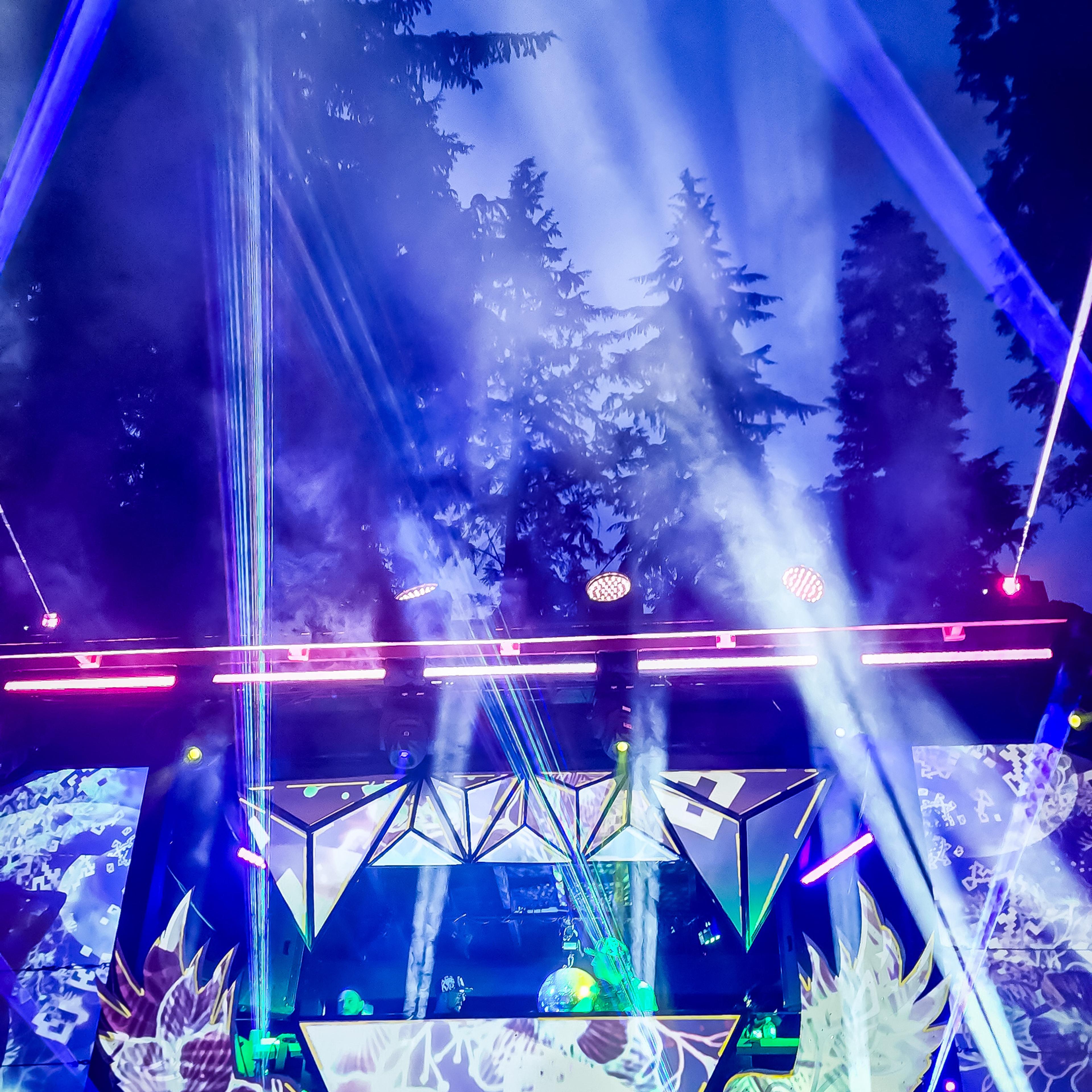 A geometric festival stage bathed in blue and purple laser beams cutting through fog above a silhouetted crowd, with tall trees lit up in the background.
