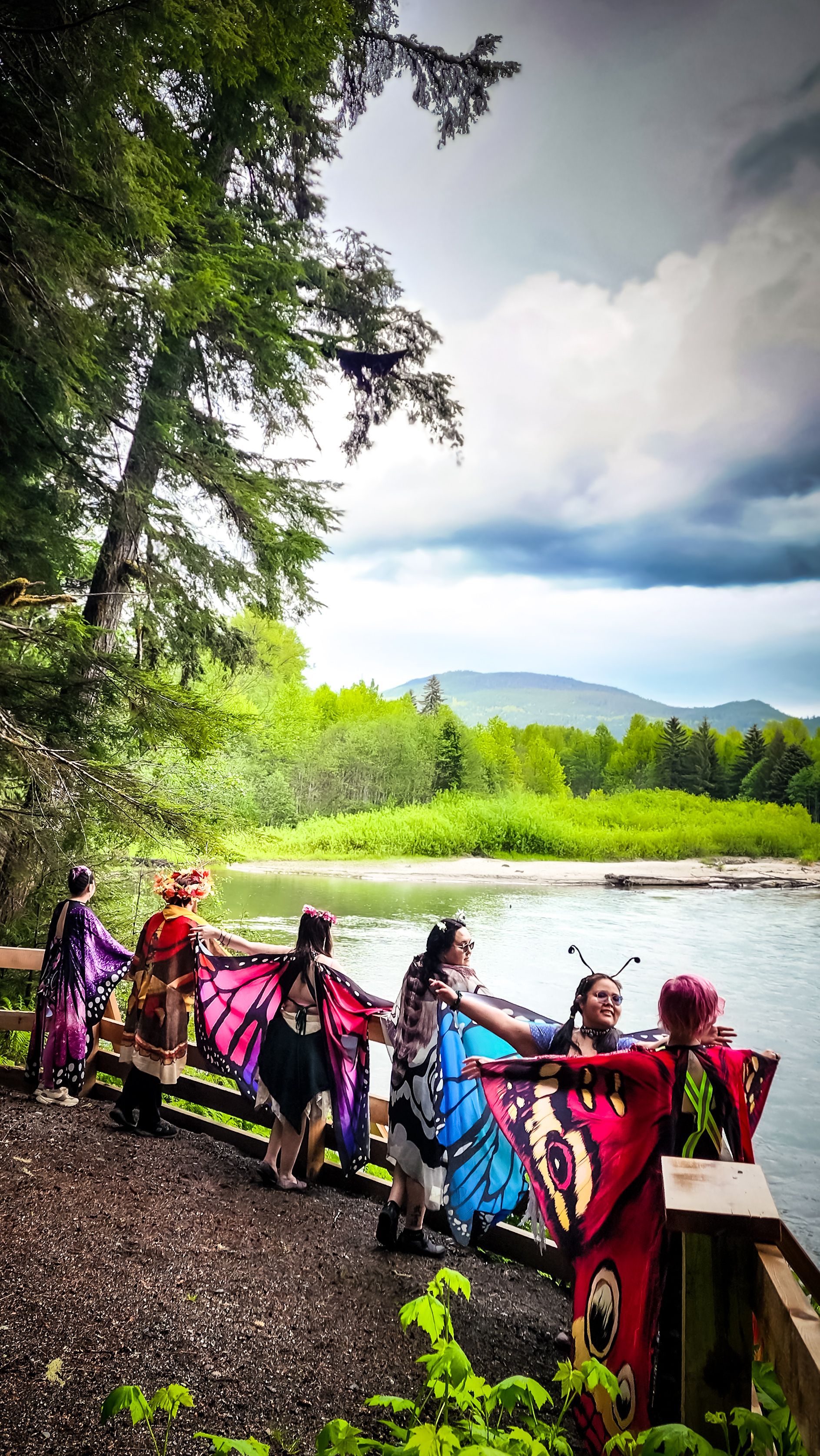 People in butterfly wings standing at a wooden railing overlooking a wide river and mountains.
