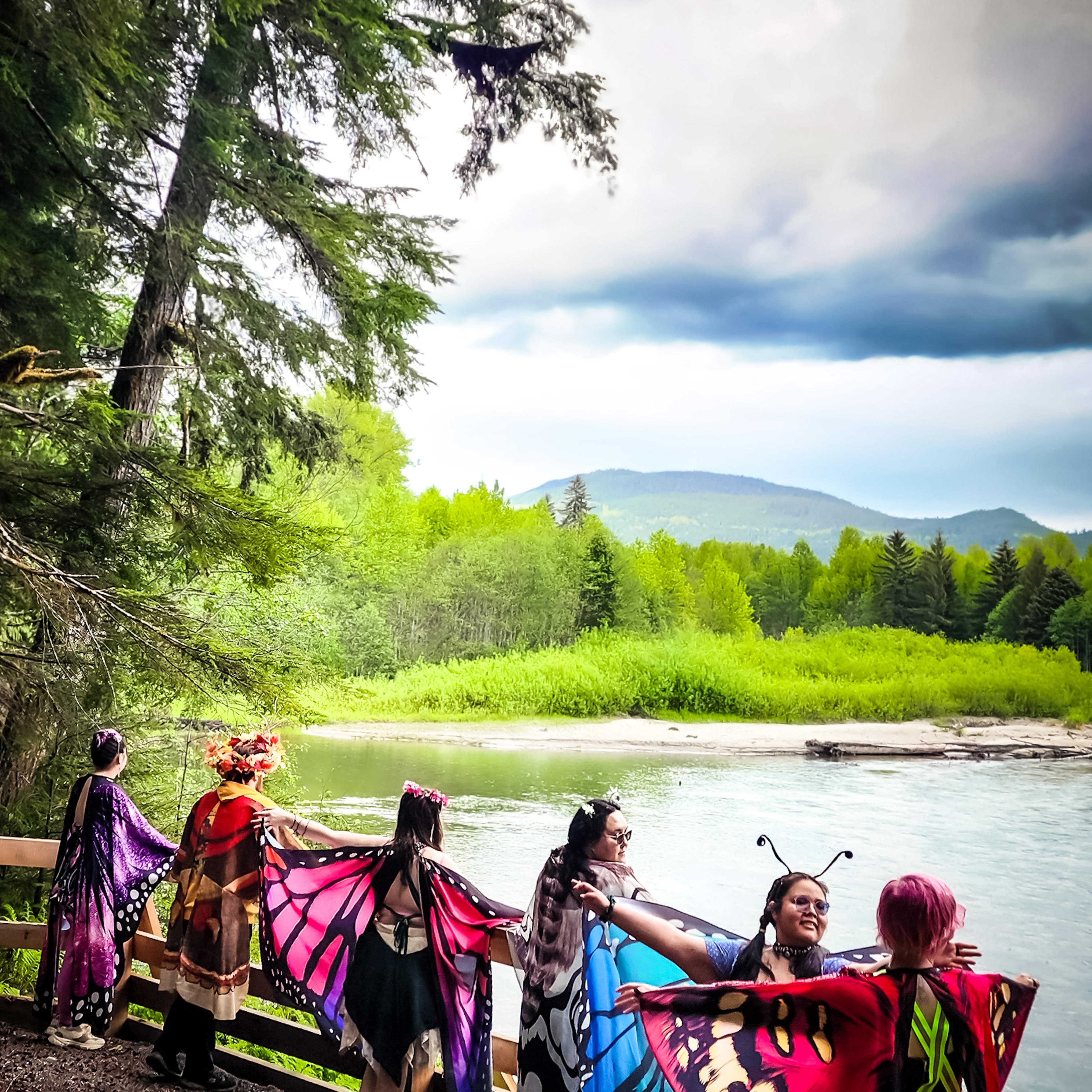 People in butterfly wings standing at a wooden railing overlooking a wide river and mountains.