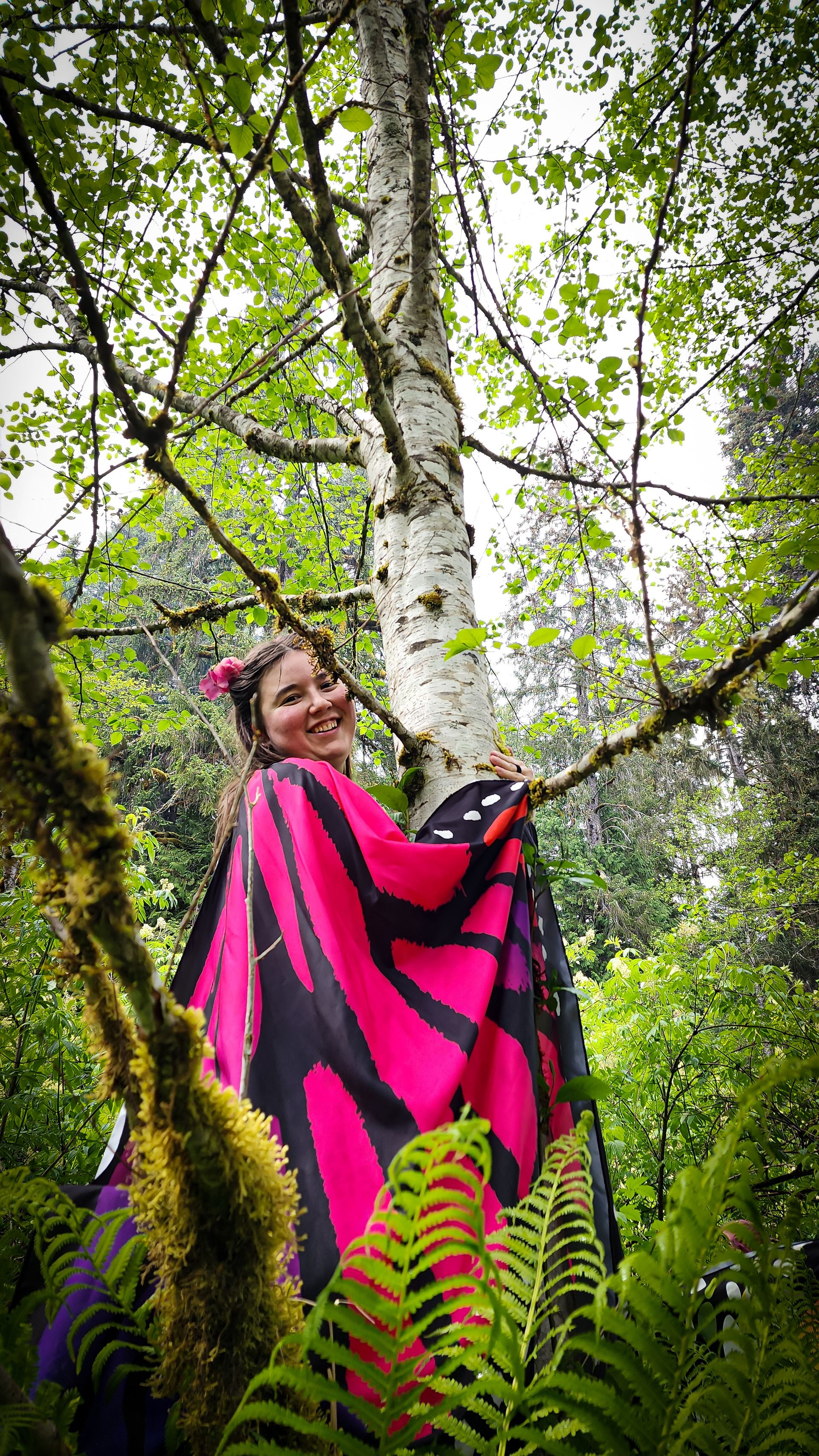 A person wearing vibrant pink butterfly wings hugging a birch tree in the forest.