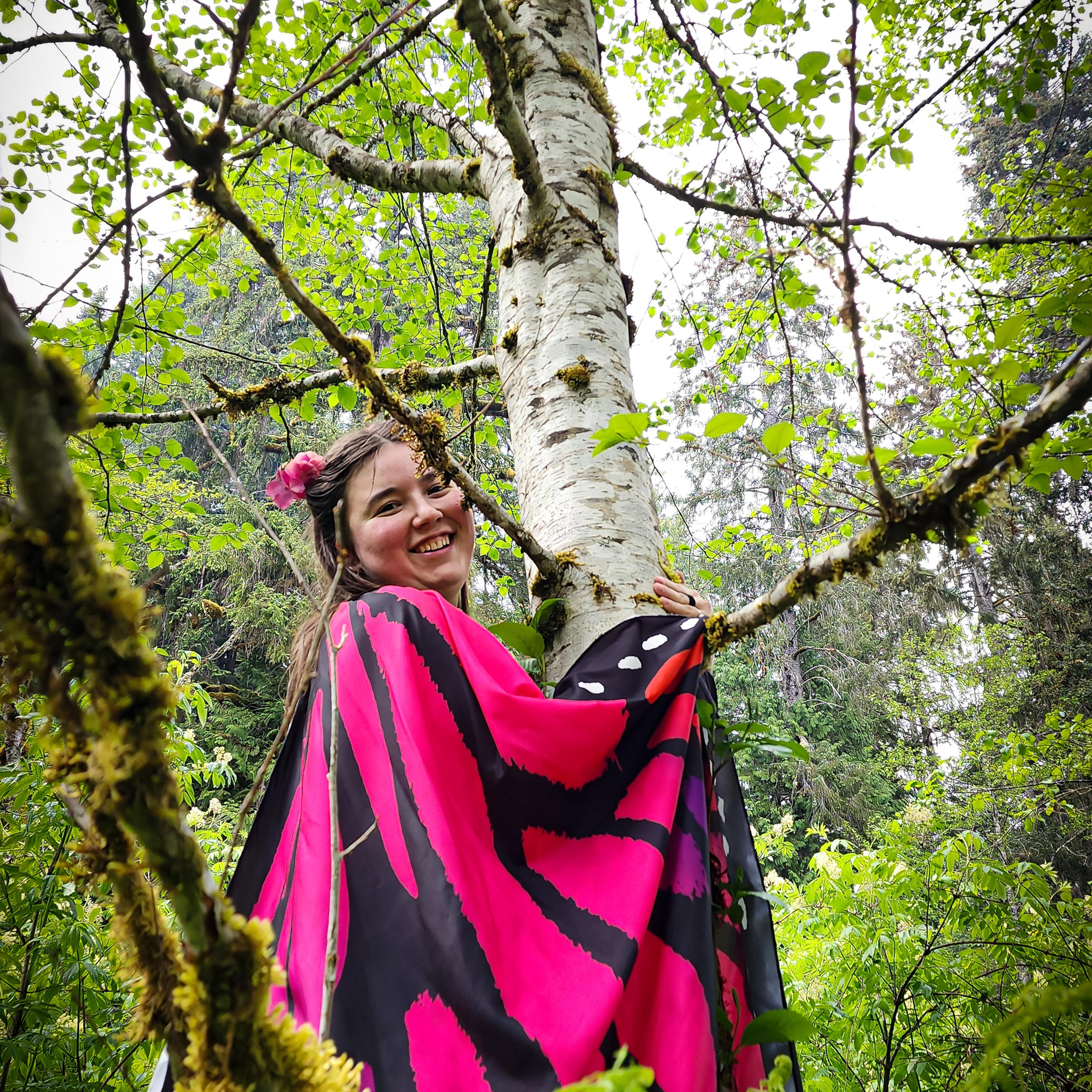 A person wearing vibrant pink butterfly wings hugging a birch tree in the forest.