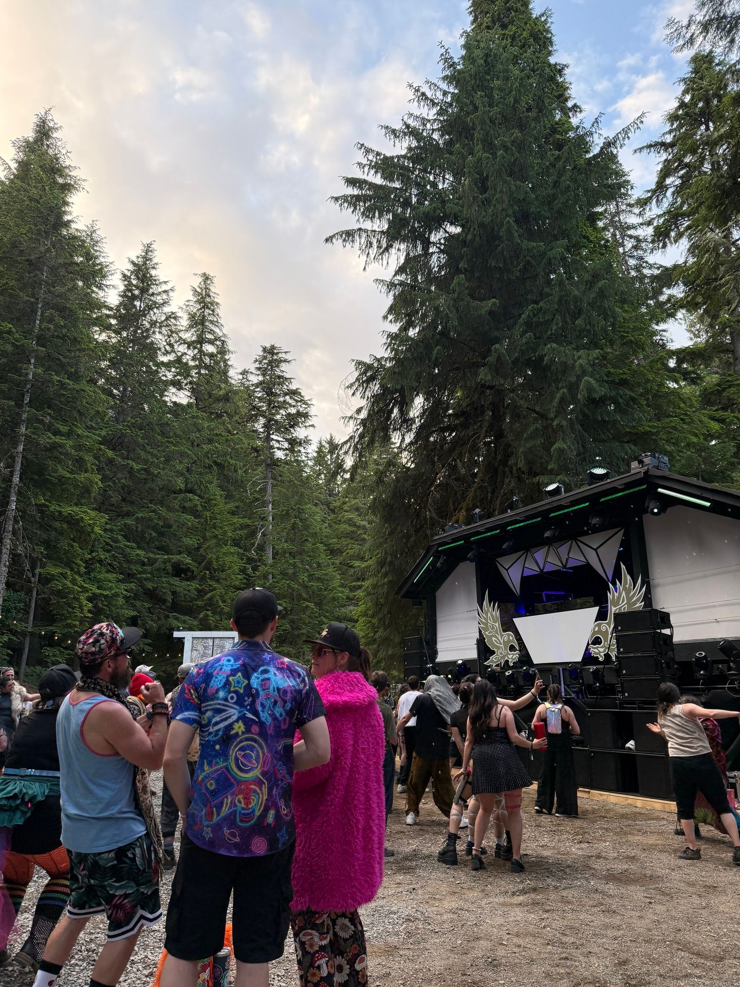 Colorfully dressed festivalgoers facing a black geometric stage nestled among tall evergreens at dusk.