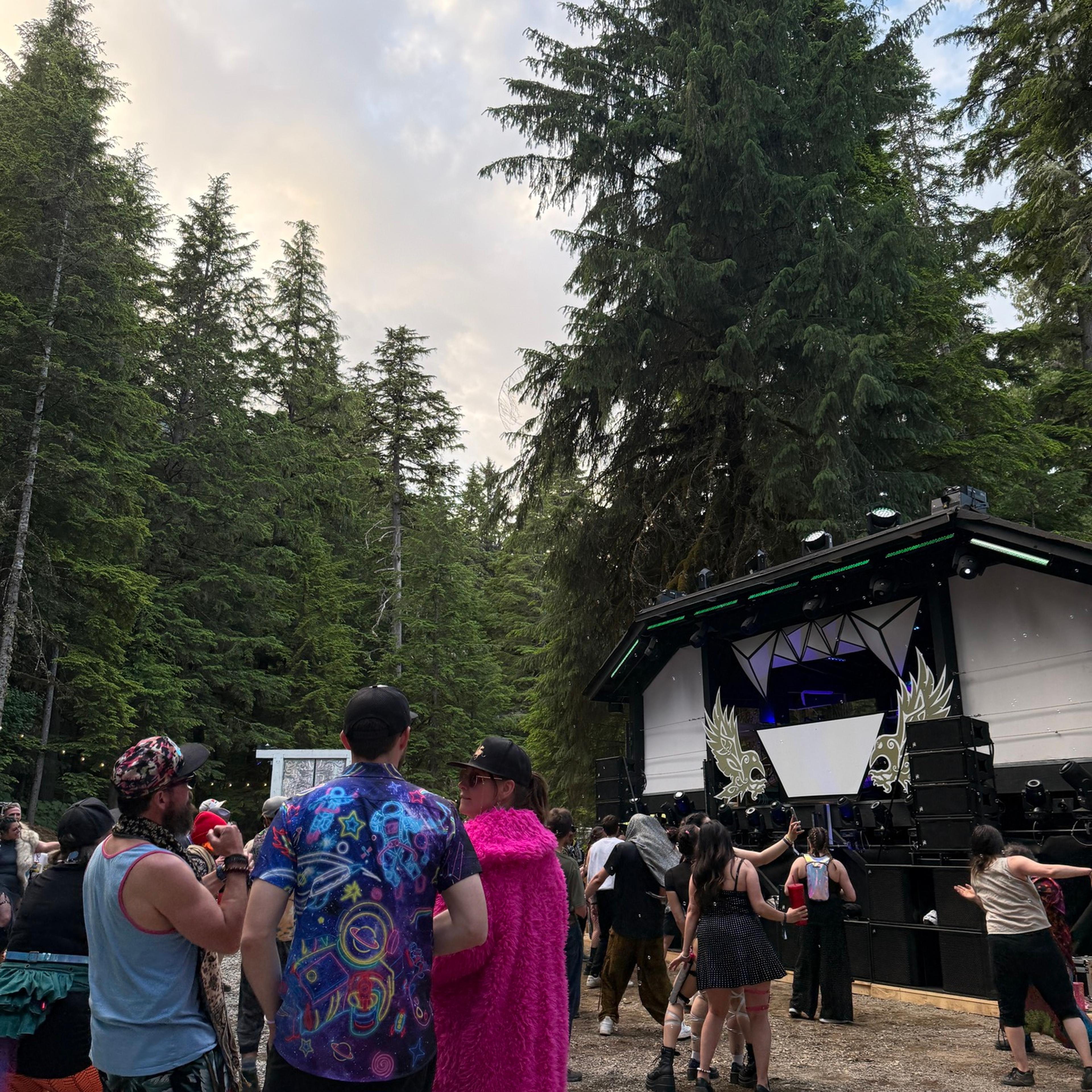 Colorfully dressed festivalgoers facing a black geometric stage nestled among tall evergreens at dusk.