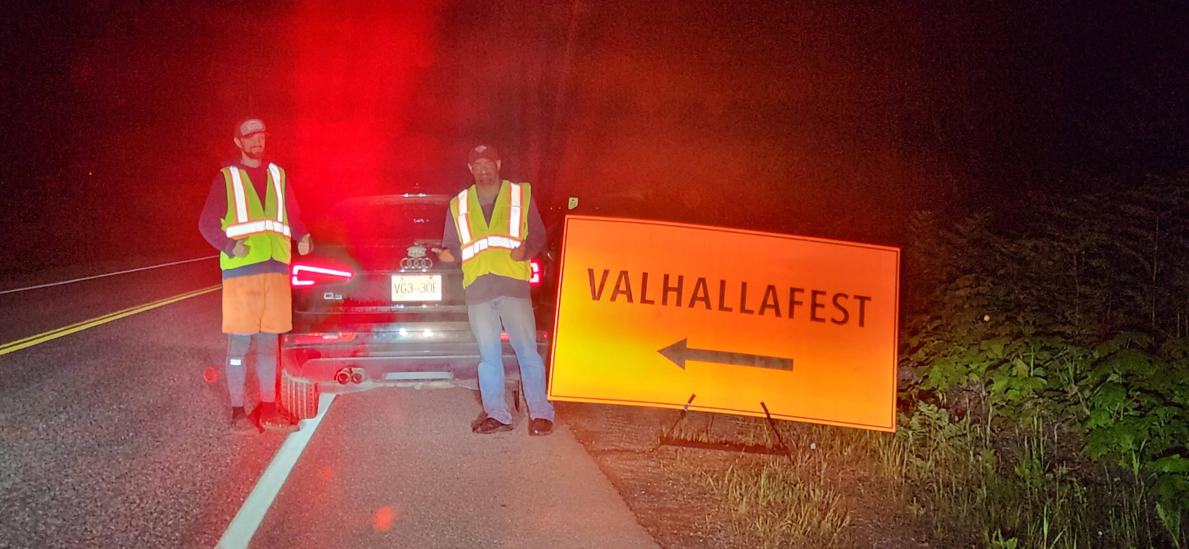 Two volunteers in high-vis vests standing beside a ValhallaFest directional sign on a dark rural road.