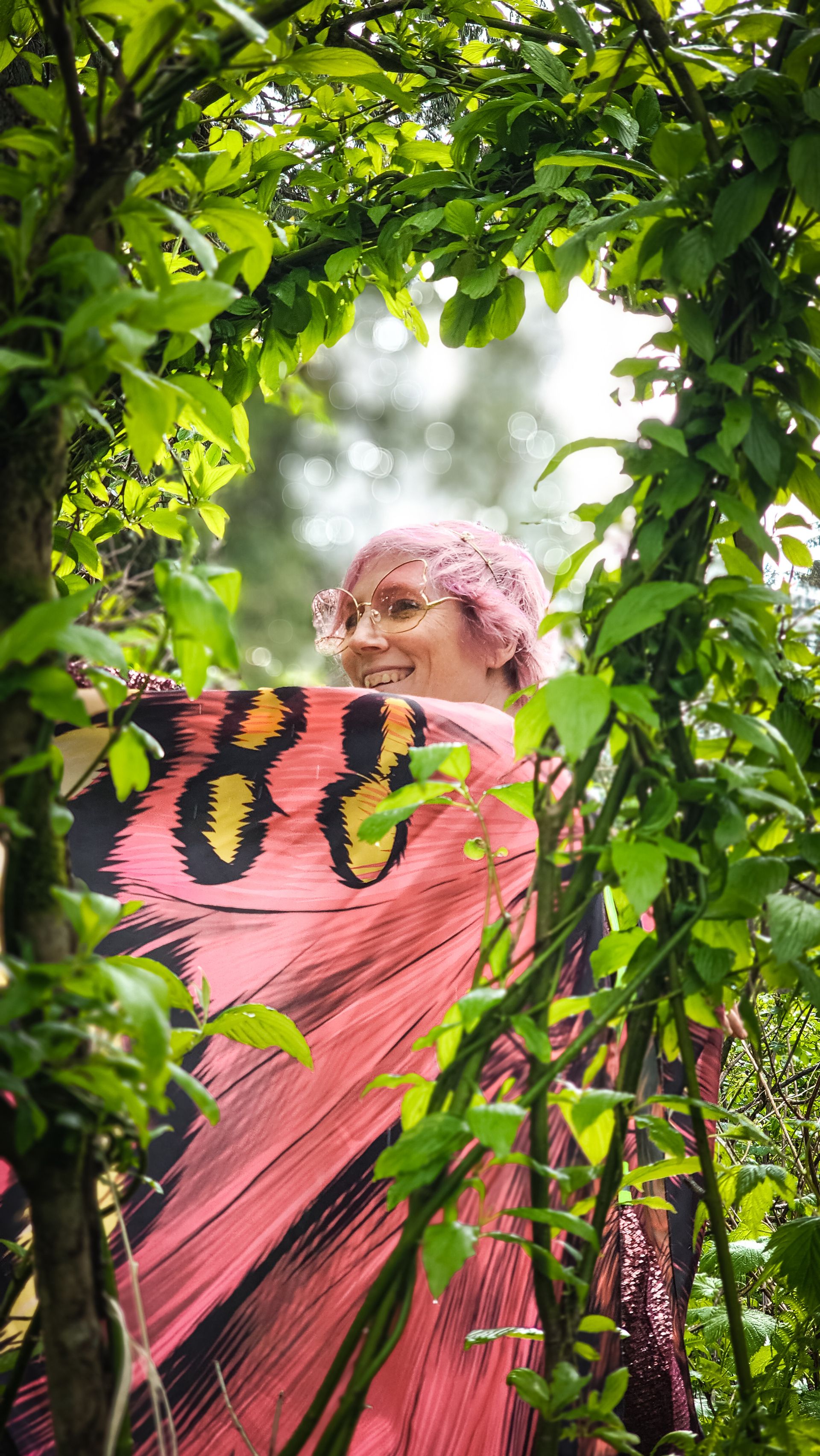 A person with pink hair and butterfly glasses smiling through a frame of green leaves.