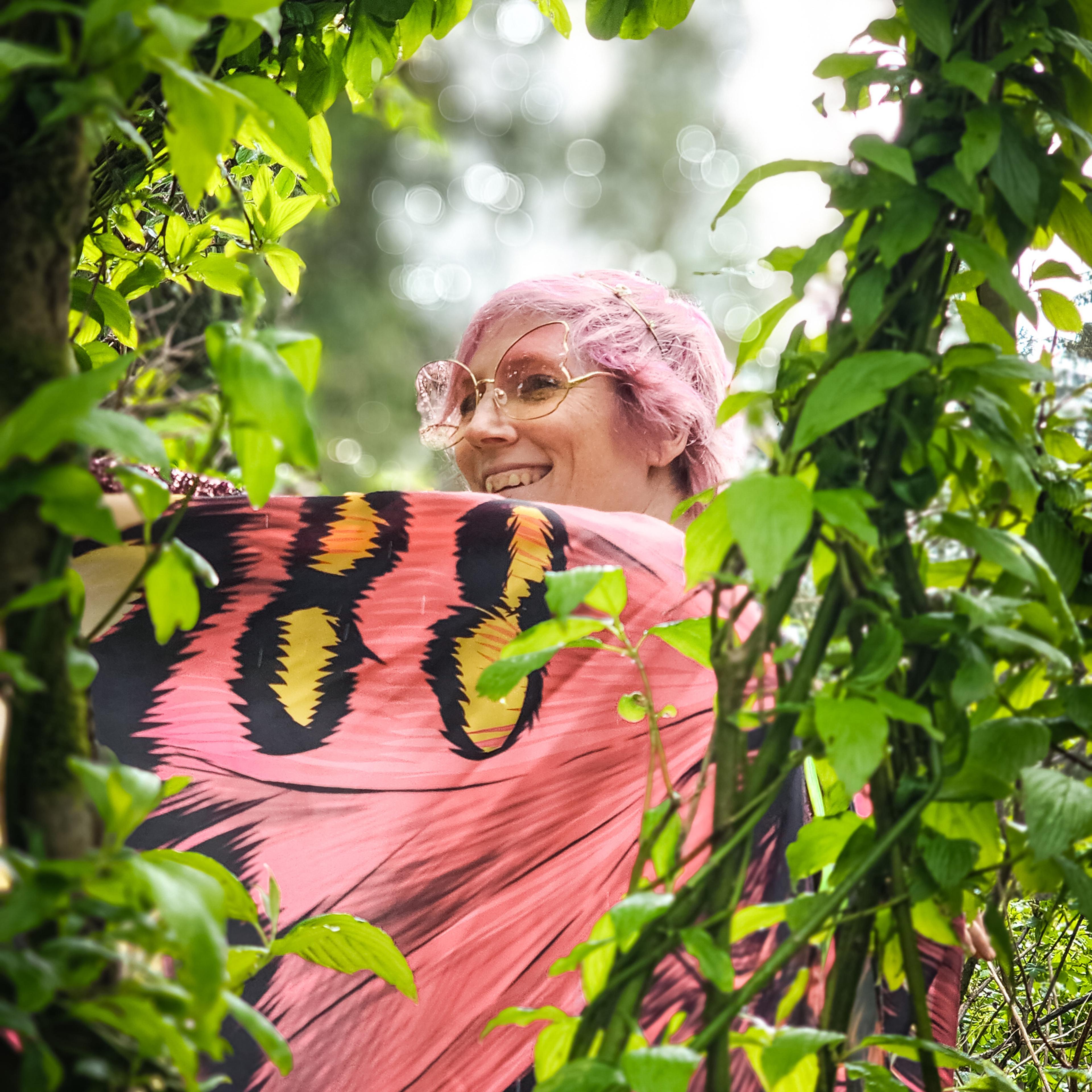 A person with pink hair and butterfly glasses smiling through a frame of green leaves.