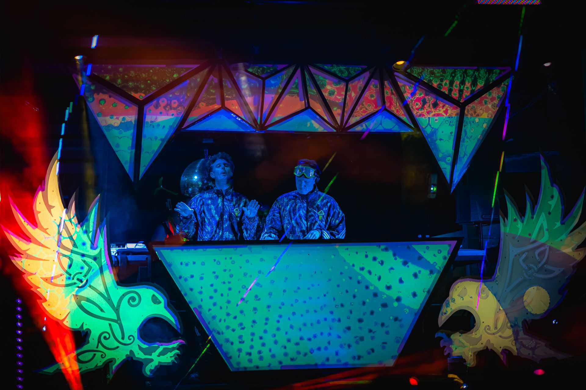 Two DJs in matching camo suits behind a glowing geometric booth, flanked by psychedelic blacklight art.