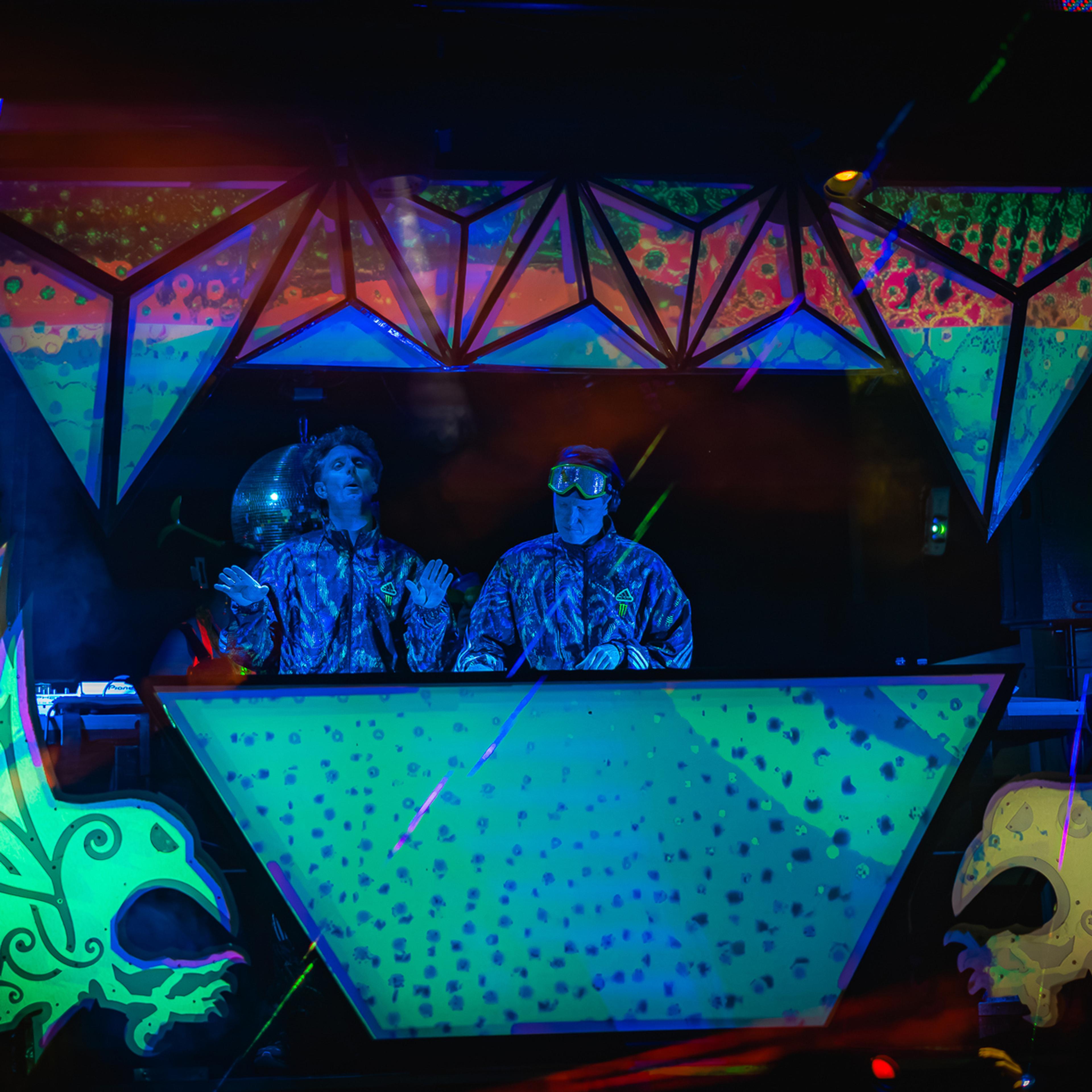 Two DJs in matching camo suits behind a glowing geometric booth, flanked by psychedelic blacklight art.