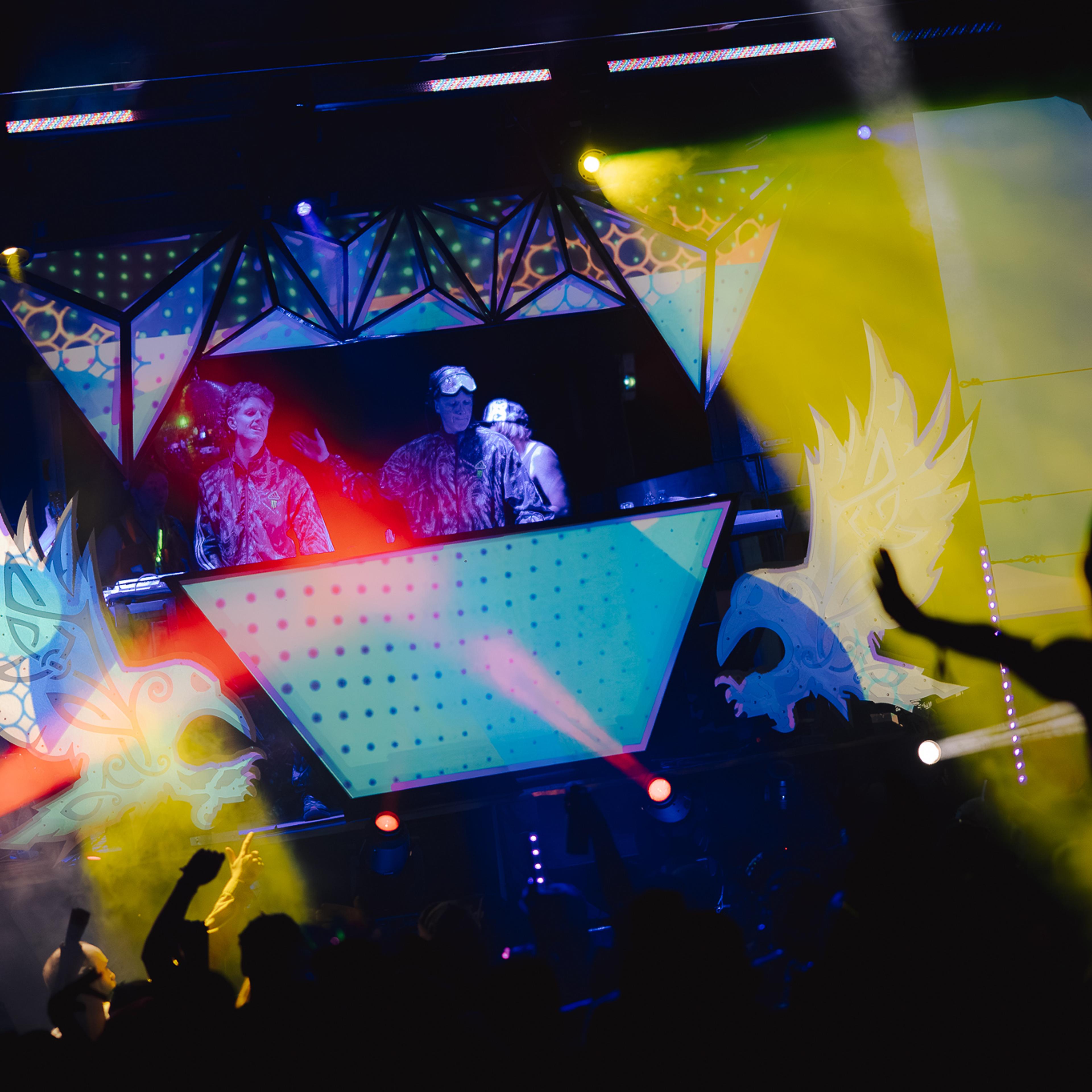 Two DJs performing on the illuminated Asgard stage with colorful lasers, smoke, and a crowd silhouetted below at ValhallaFest