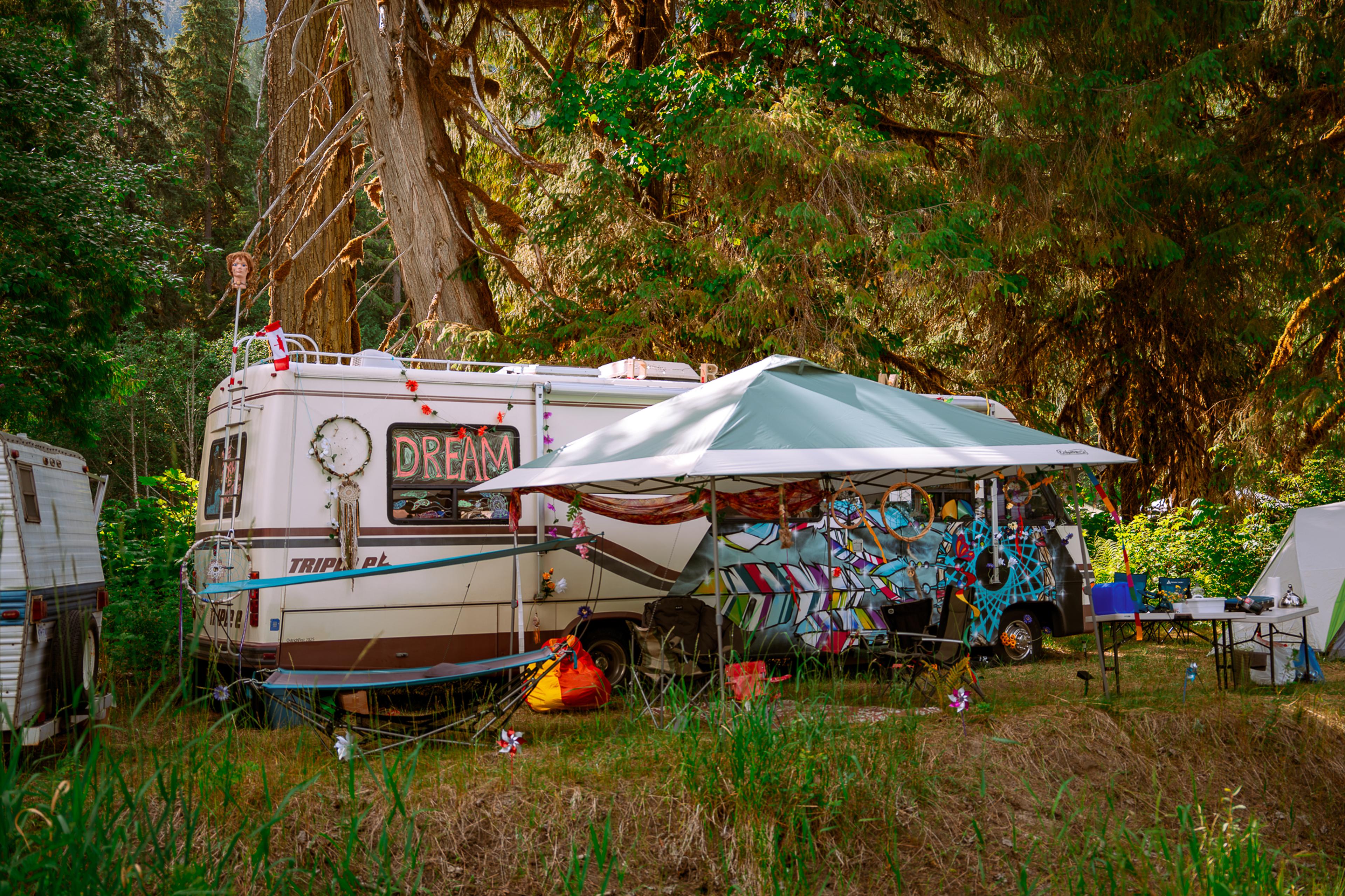 Decorated RV with a 'Dream' sign and dreamcatchers parked beside a colorfully painted vehicle under tall forest trees at ValhallaFest