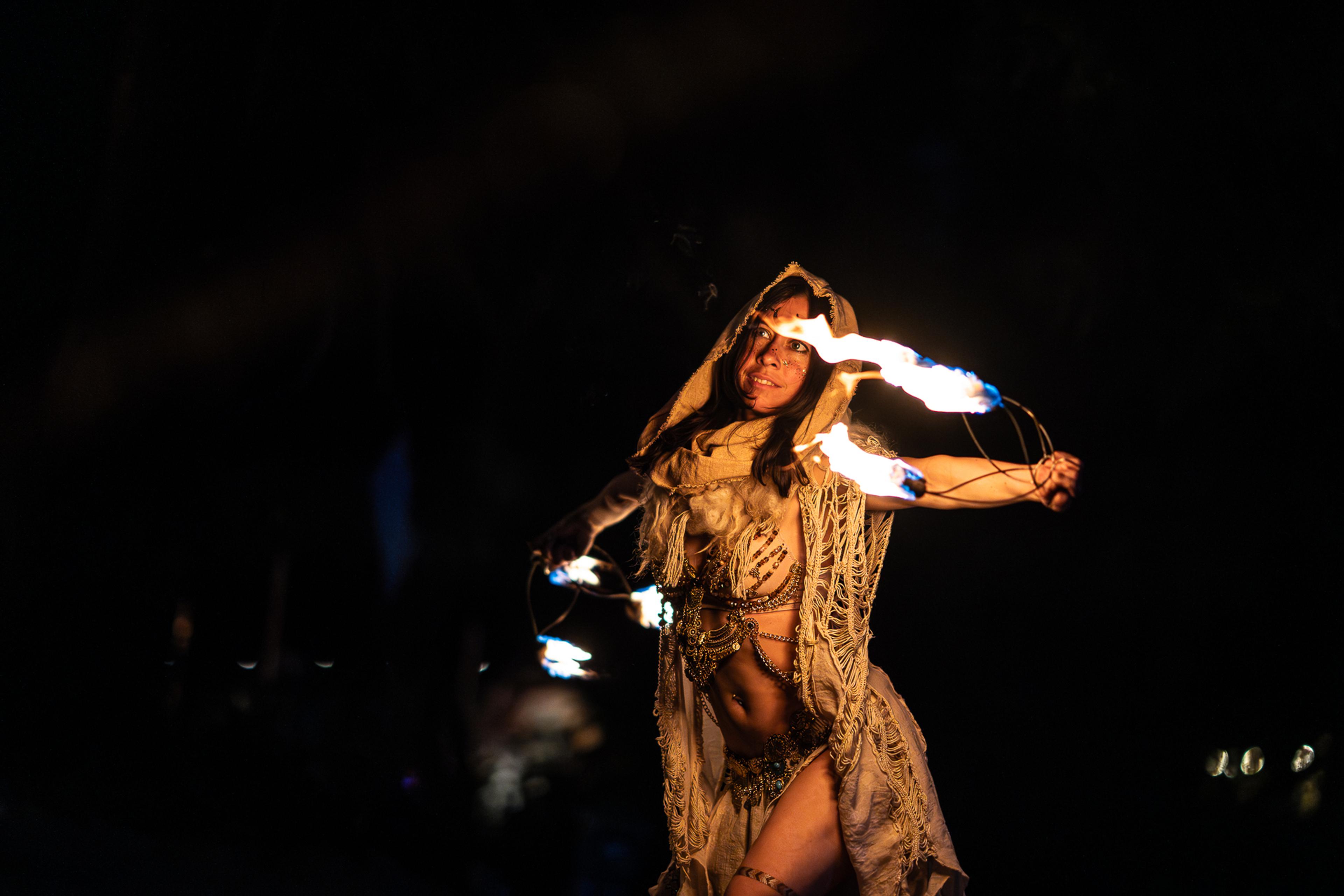 A fire performer wearing a hooded cream outfit and intricate jewelry spins flaming props against a black background.