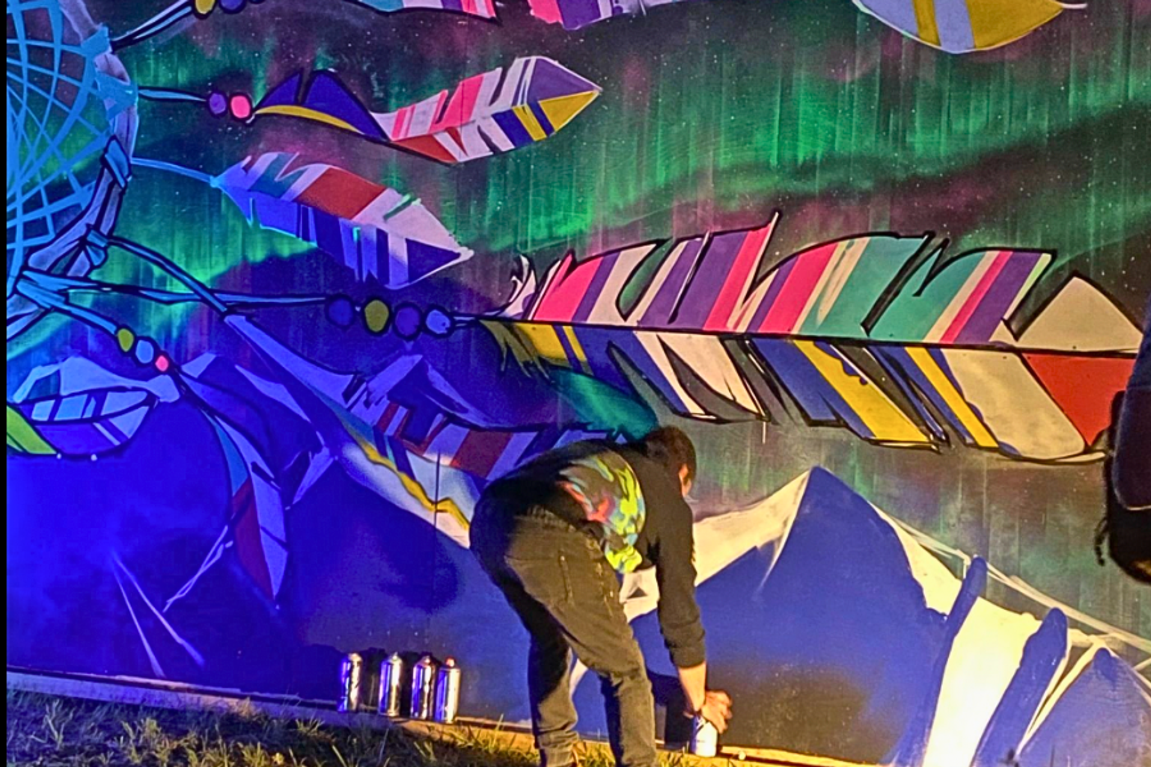 Artist spray painting a vibrant mural of feathers and northern lights at night at ValhallaFest