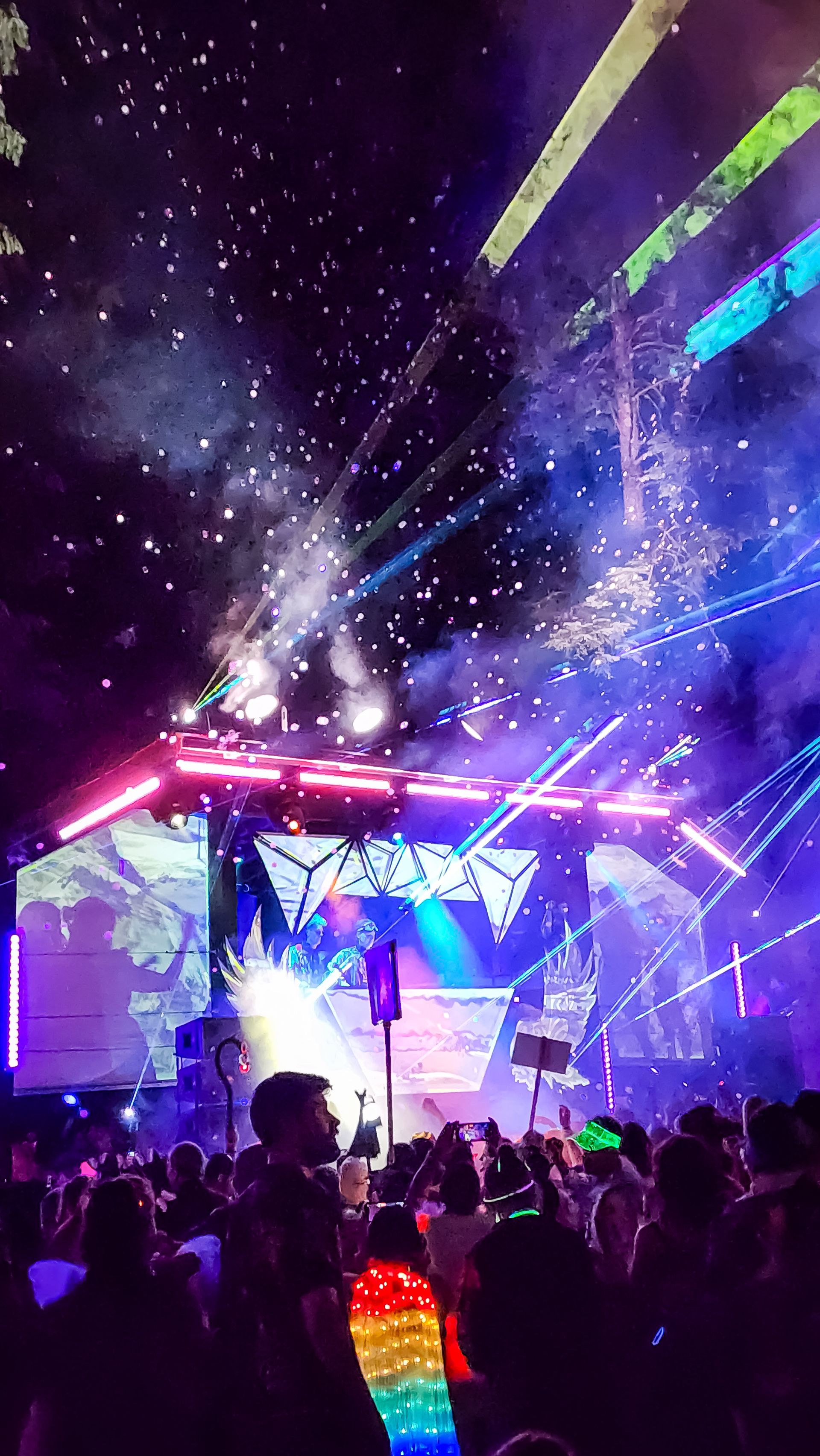 Crowd dancing under a shower of bubbles with rainbow laser beams cutting through a foggy night sky above the stage.