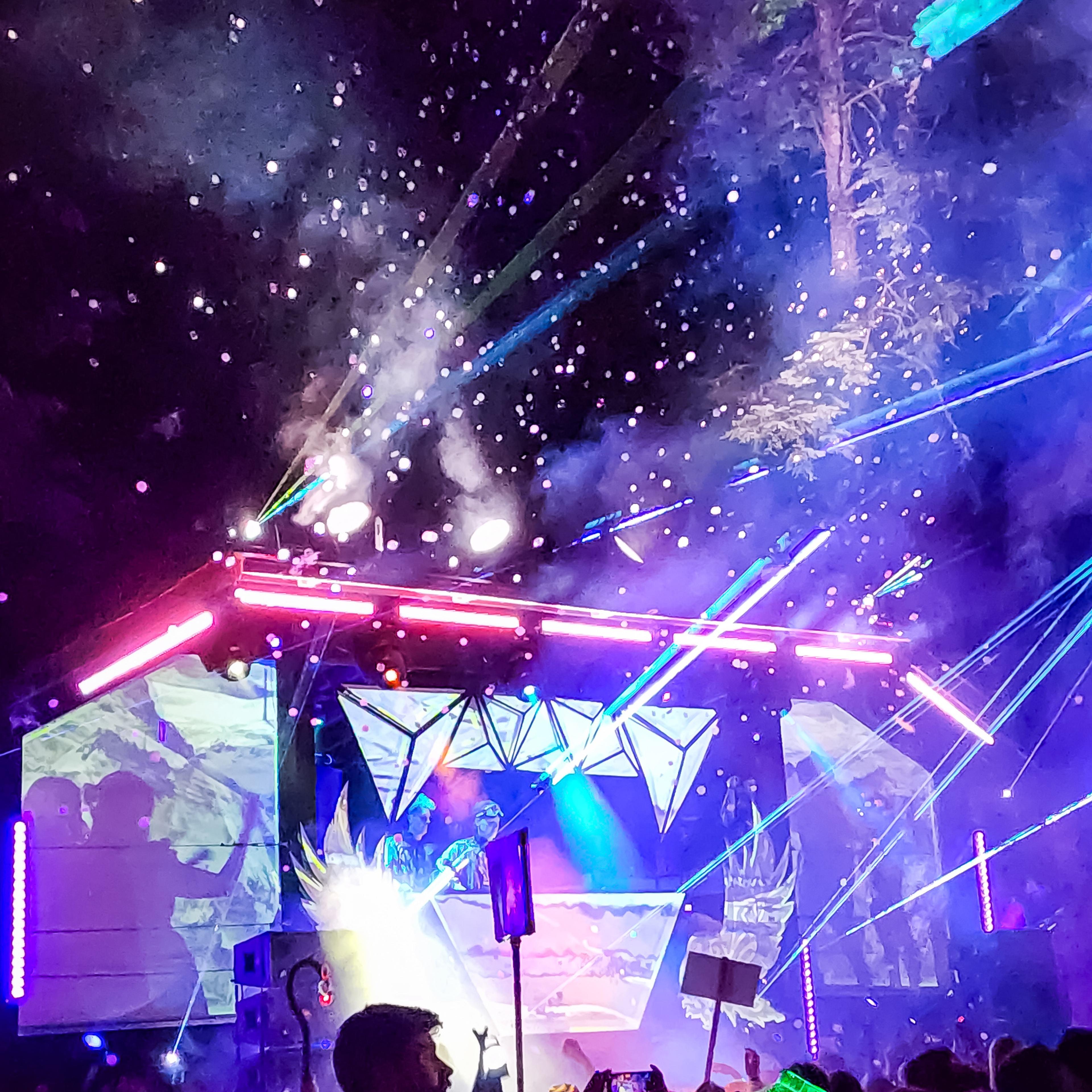 Crowd dancing under a shower of bubbles with rainbow laser beams cutting through a foggy night sky above the stage.