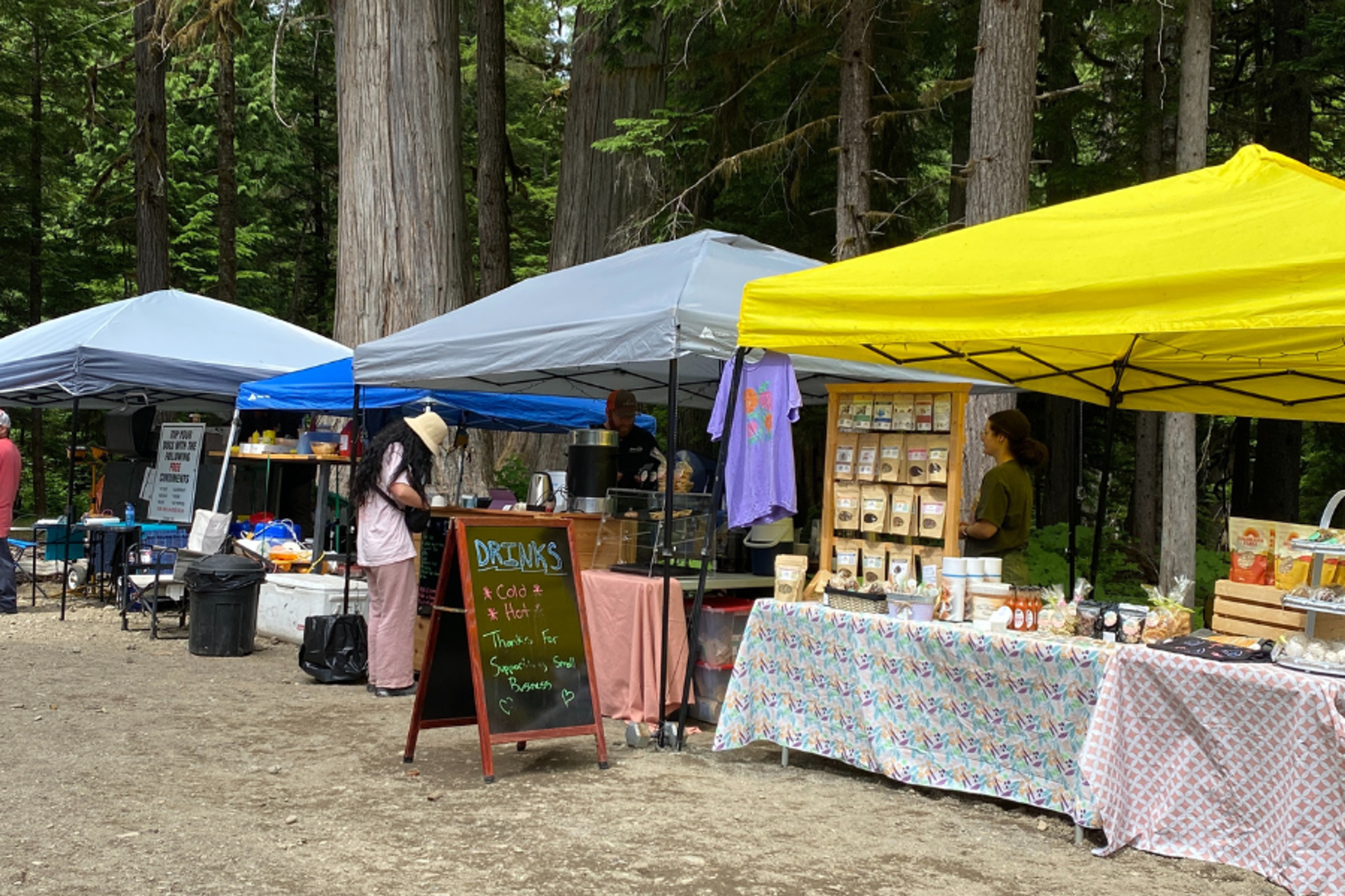 Outdoor market stalls selling drinks and goods beneath canopy tents in a forested festival area at Valhallafest