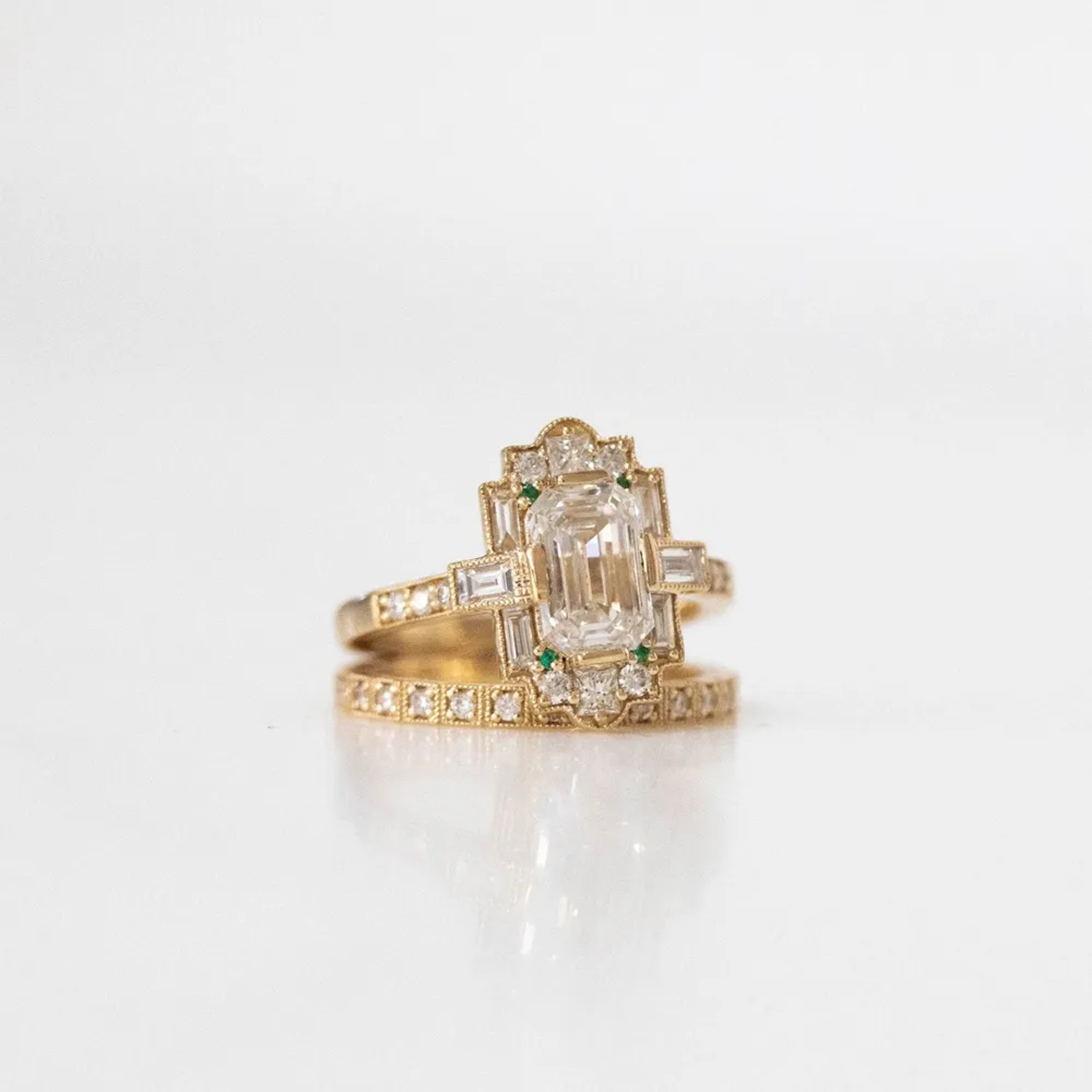 a close-up of a yellow gold, emerald-cut diamond ring with a halo of smaller emerald-cut diamonds, sitting on top of a wedding band against a plain white background