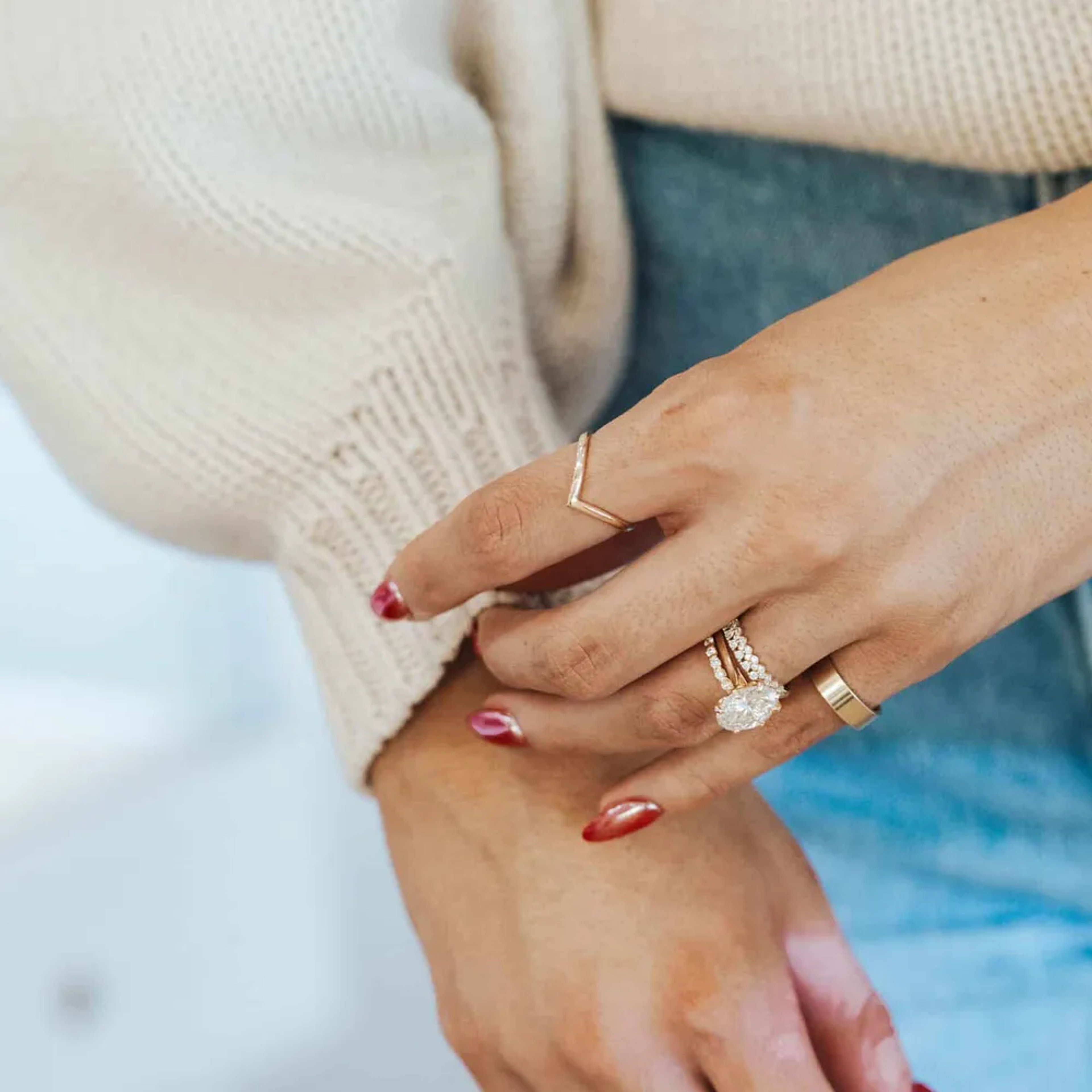 A close-up of a woman's hands, wearing a very large diamond ring on her left hand. The woman is wearing a cream sweater and has red nail polish on her fingers.