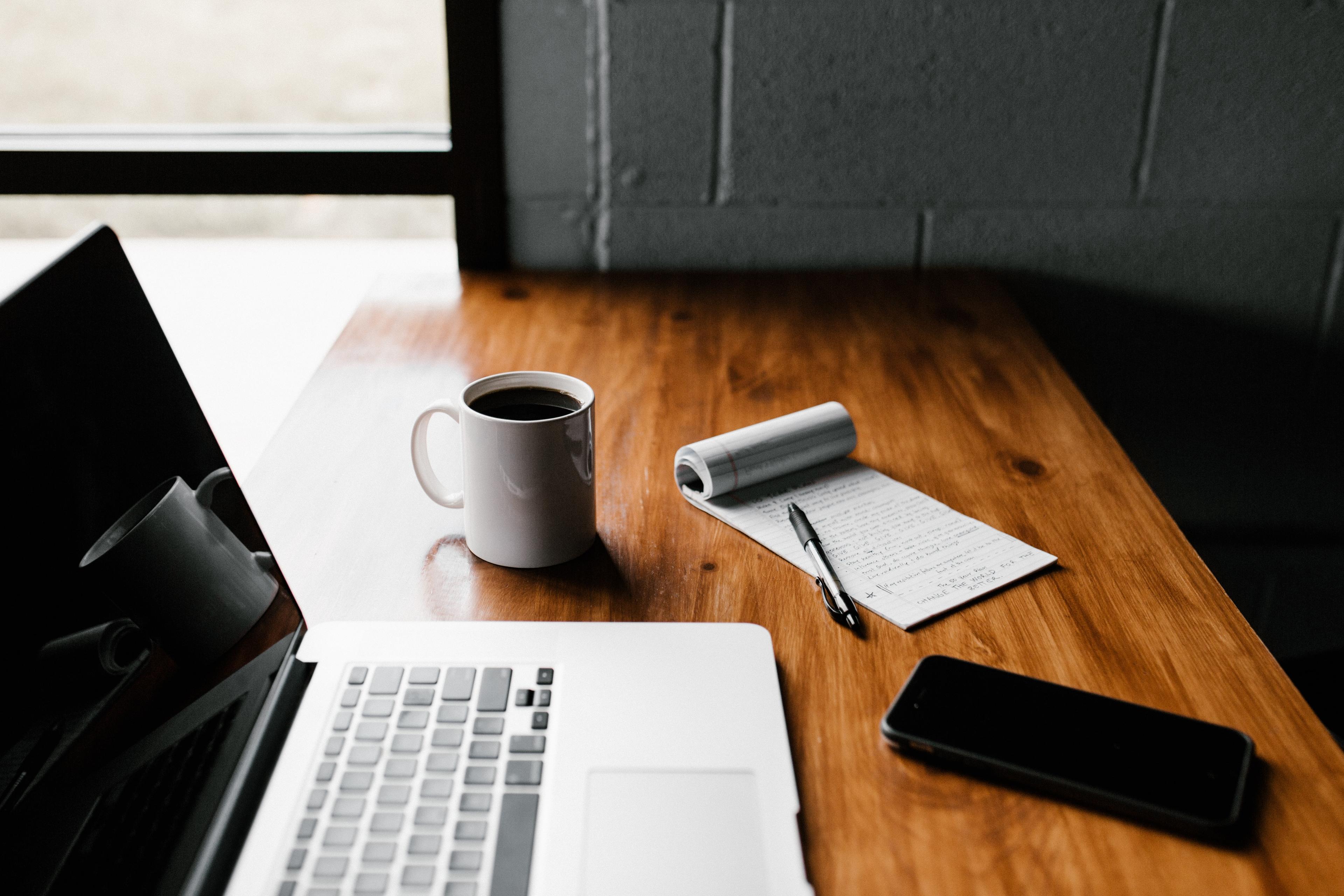 A laptop, a cup filled with coffee, a notebook, a pen, and a phone are kept on a desk. 