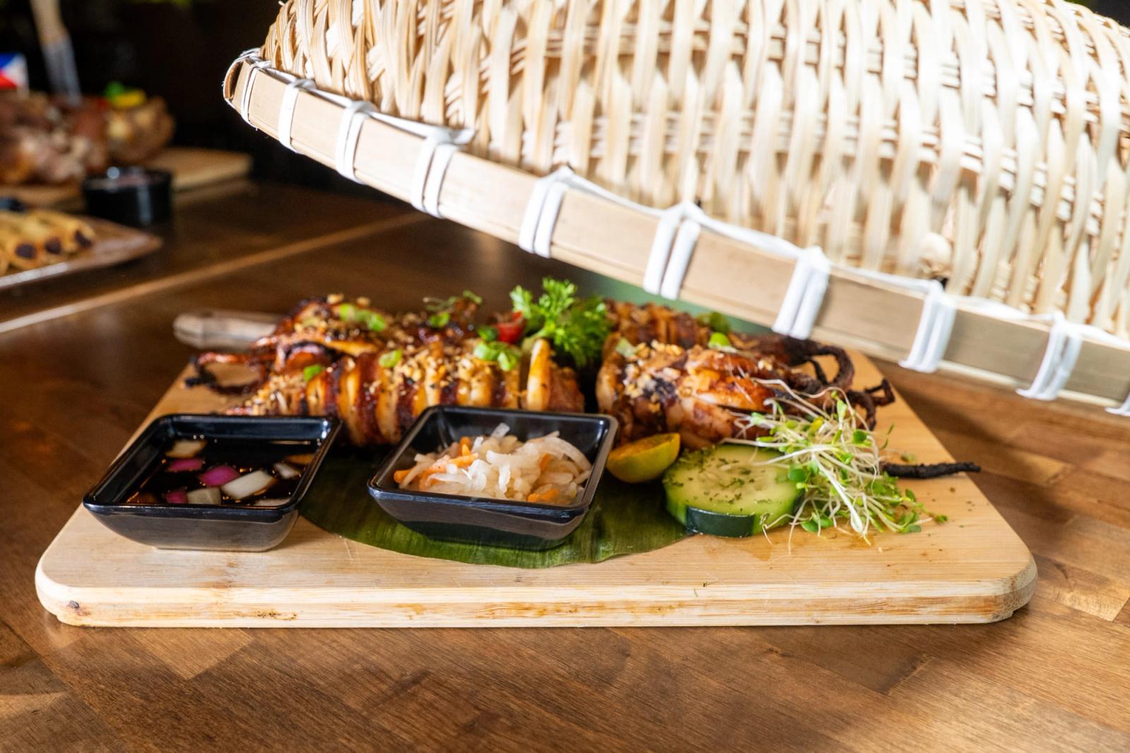 A wicker cover lifts off a wooden board of grilled skewers with dipping sauces, cucumber slices and microgreens on a restaurant table.