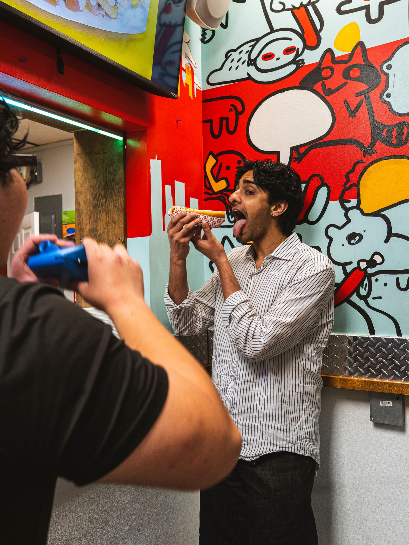 A crew member with a camera photographs a man playfully sticking out his tongue at a hot dog in front of a colorful cartoon mural.