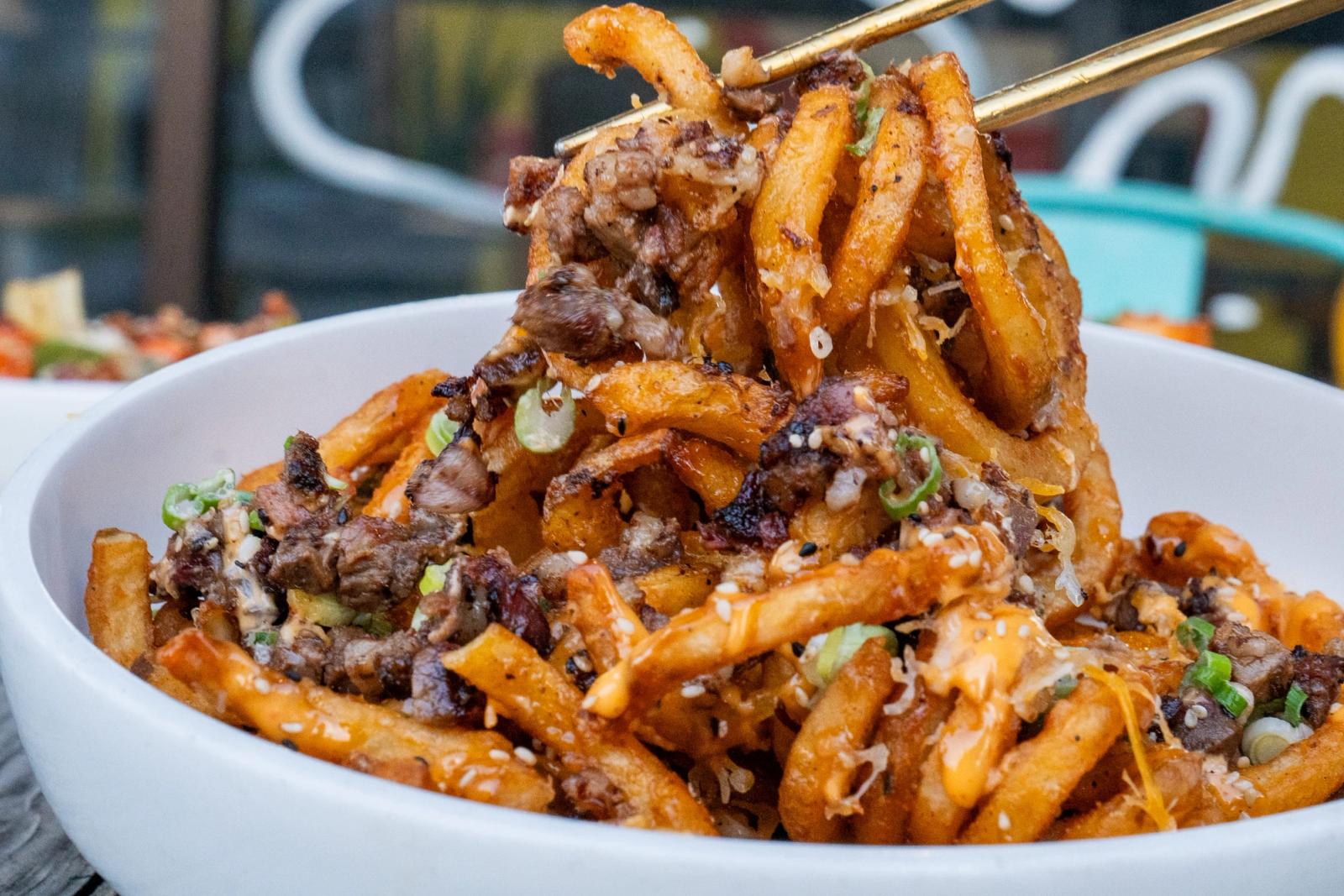 Chopsticks lift loaded fries topped with seasoned ground meat and green onions from a white bowl on an outdoor table.