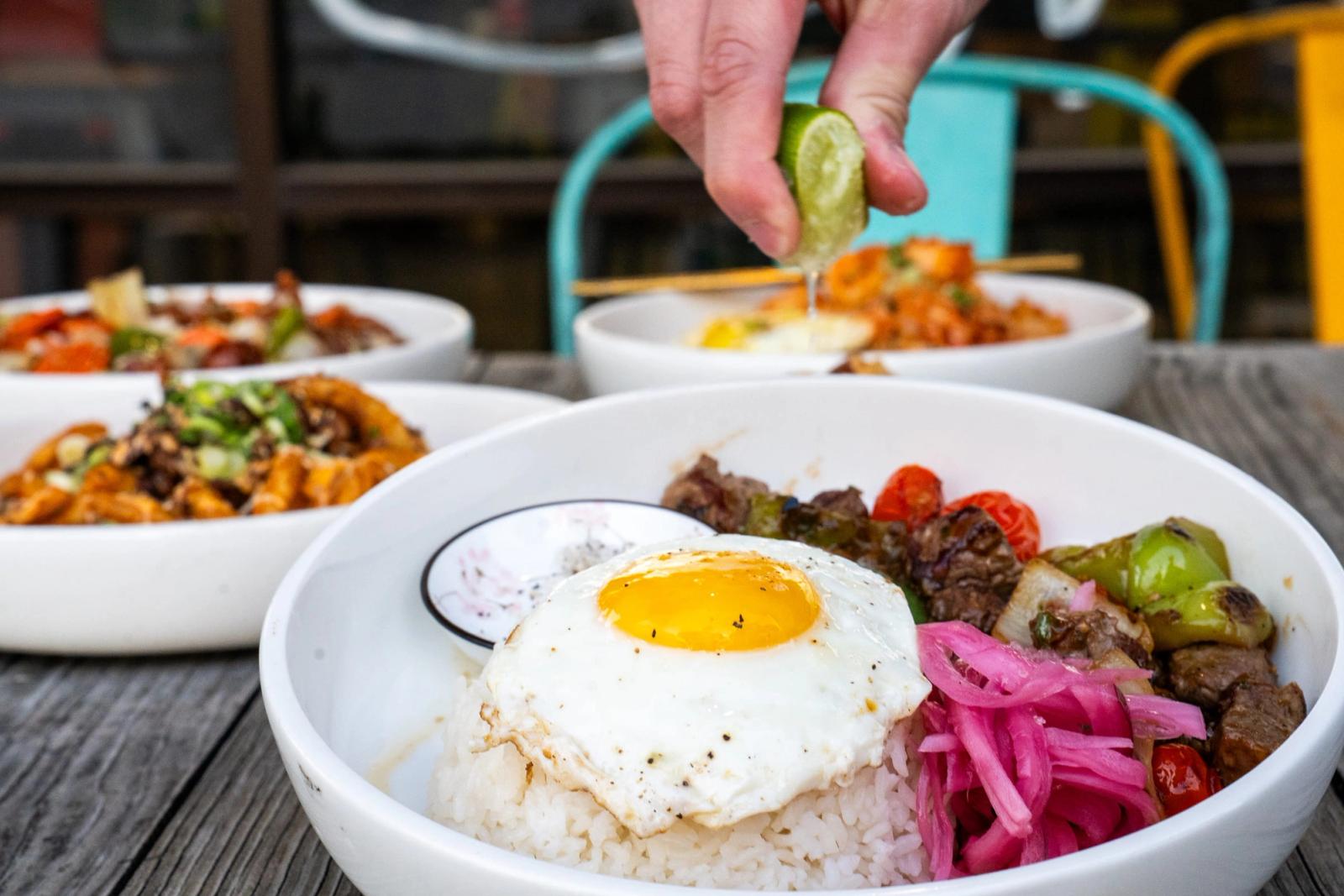 A hand squeezes lime over a rice bowl with a fried egg, pickled onion and beef, with other bowls on an outdoor patio table.