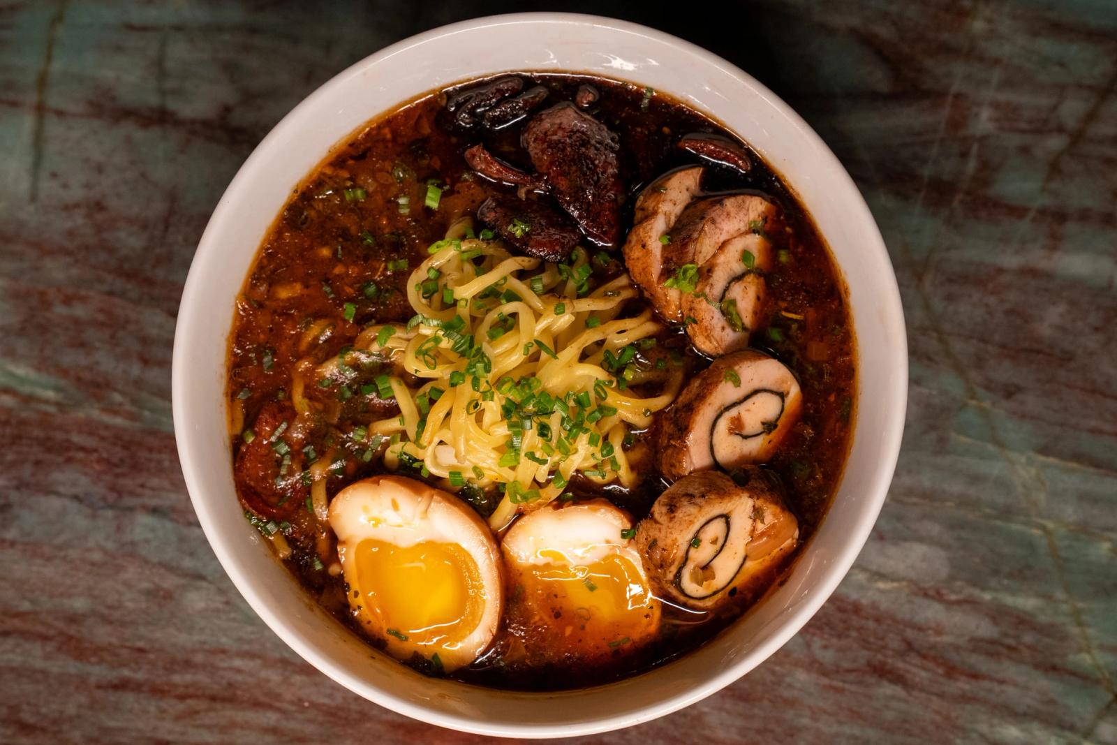 Overhead view of a bowl of dark-broth ramen with chashu pork, halved soft-boiled eggs, noodles, scallions and mushrooms on a marble table.