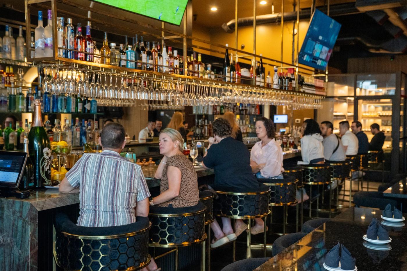 Guests sit on gold-trimmed stools at a busy bar in front of a lit back bar with bottles, hanging glassware and TVs.