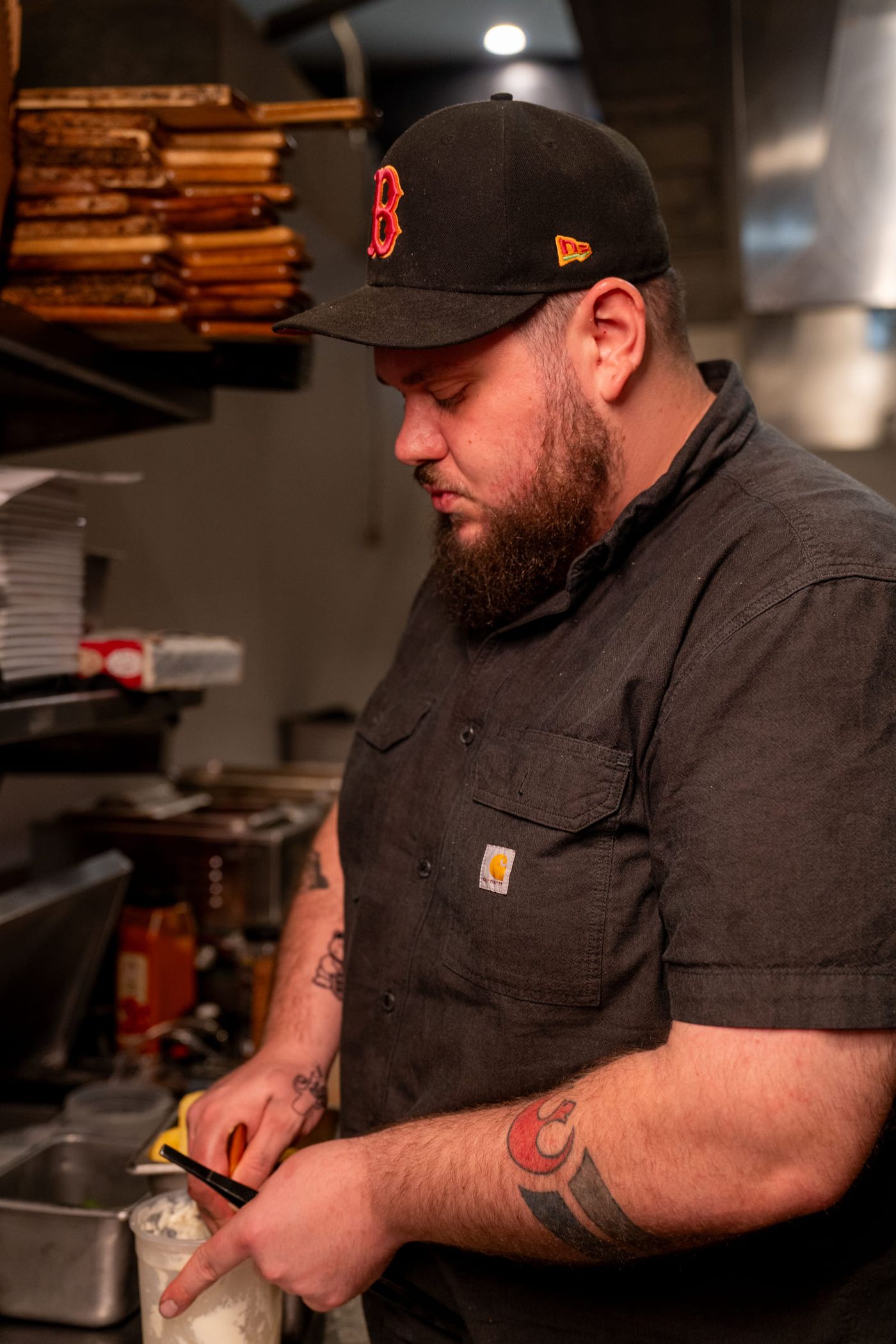 A bearded cook in a baseball cap and work shirt preps food at a kitchen station.