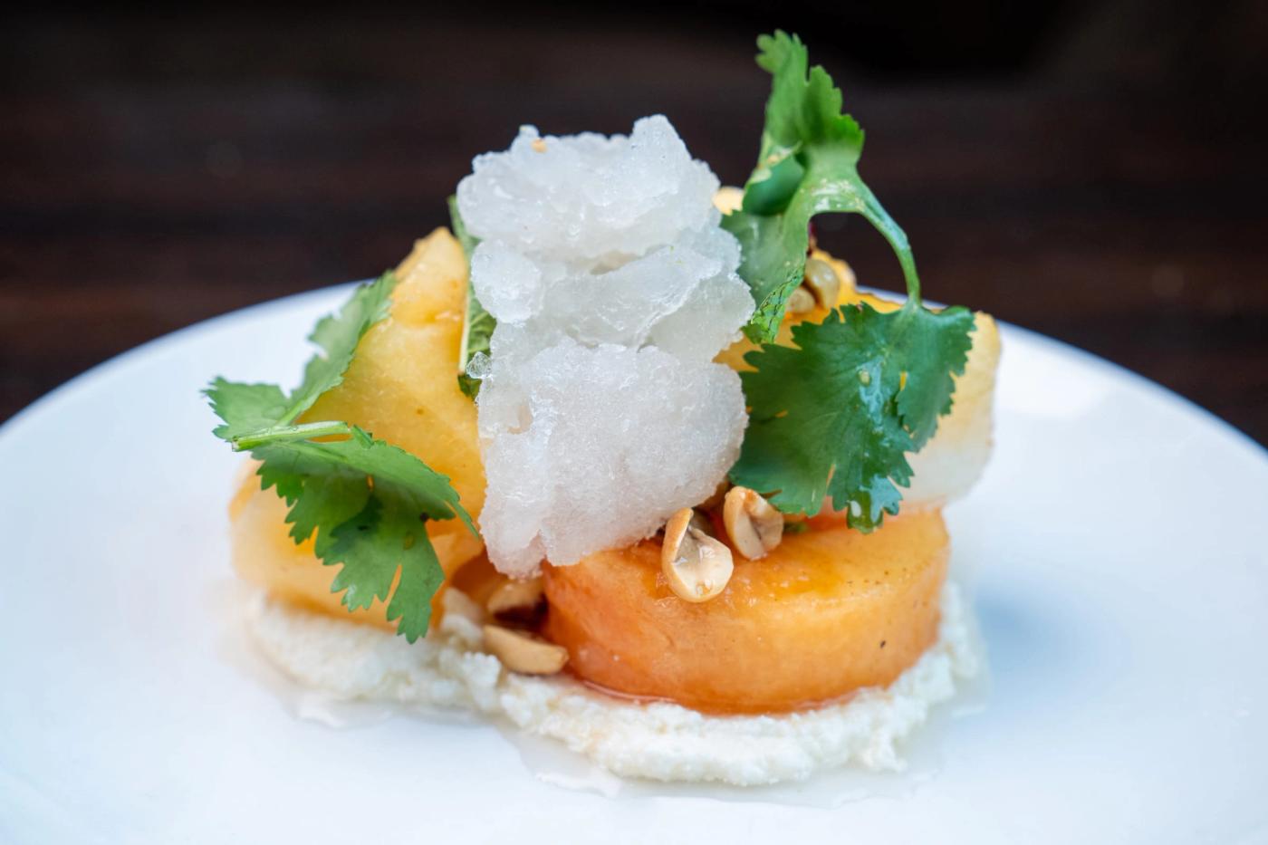 Sliced orange melon topped with shaved granita, cilantro and peanuts on a cream base, served on a white plate.