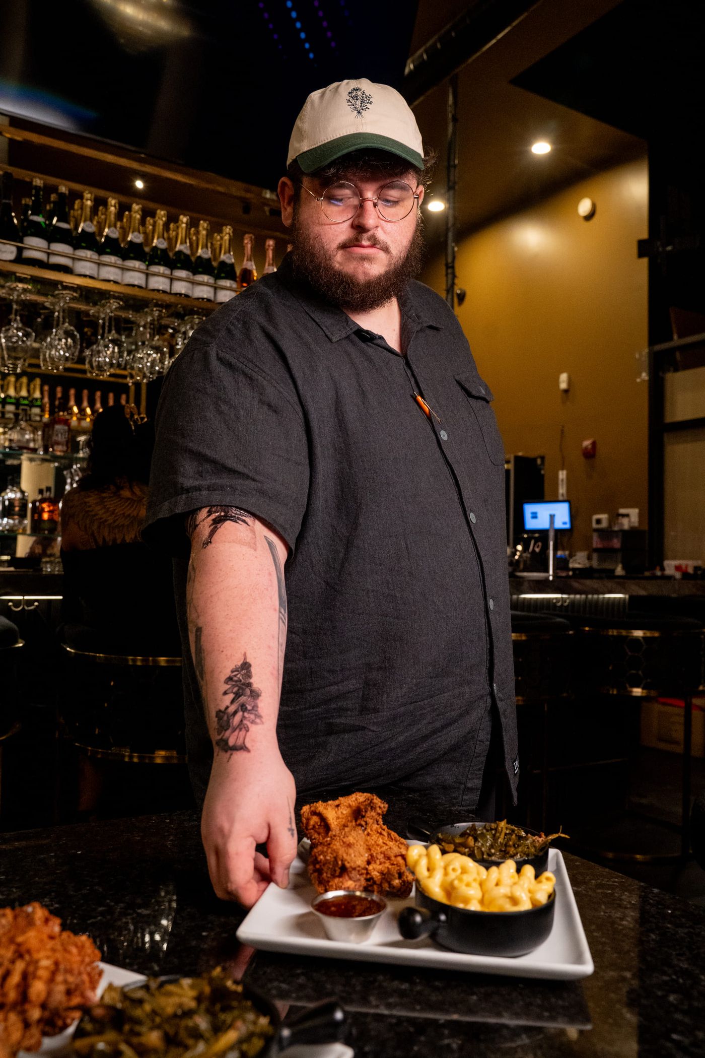 A bearded cook in a cap and glasses sets down a platter of fried chicken, mac and cheese and greens on a bar counter.