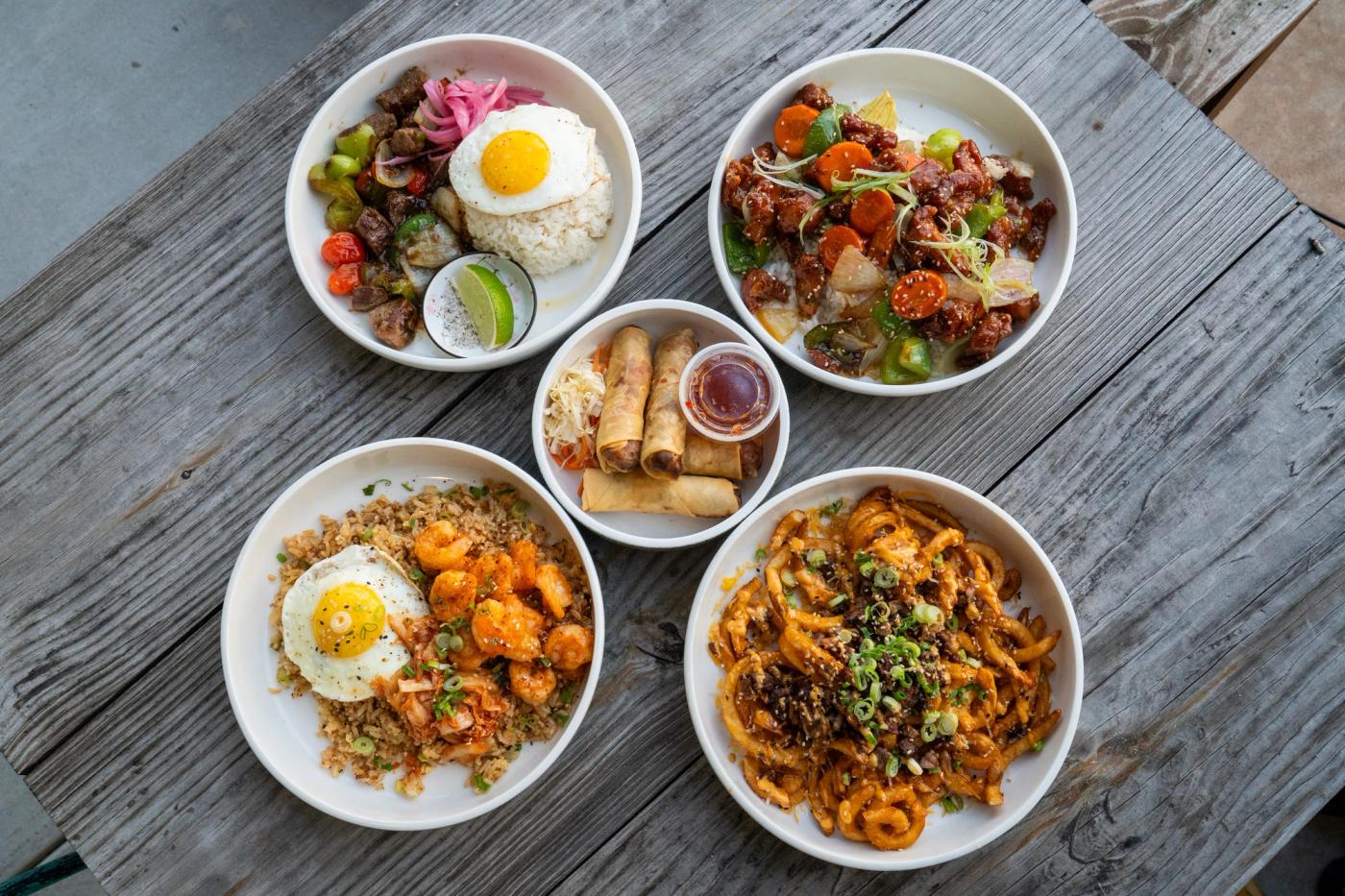 Five dishes seen from above on a wooden table, including a rice plate with egg, egg rolls with sauce, shrimp fried rice and loaded fries.