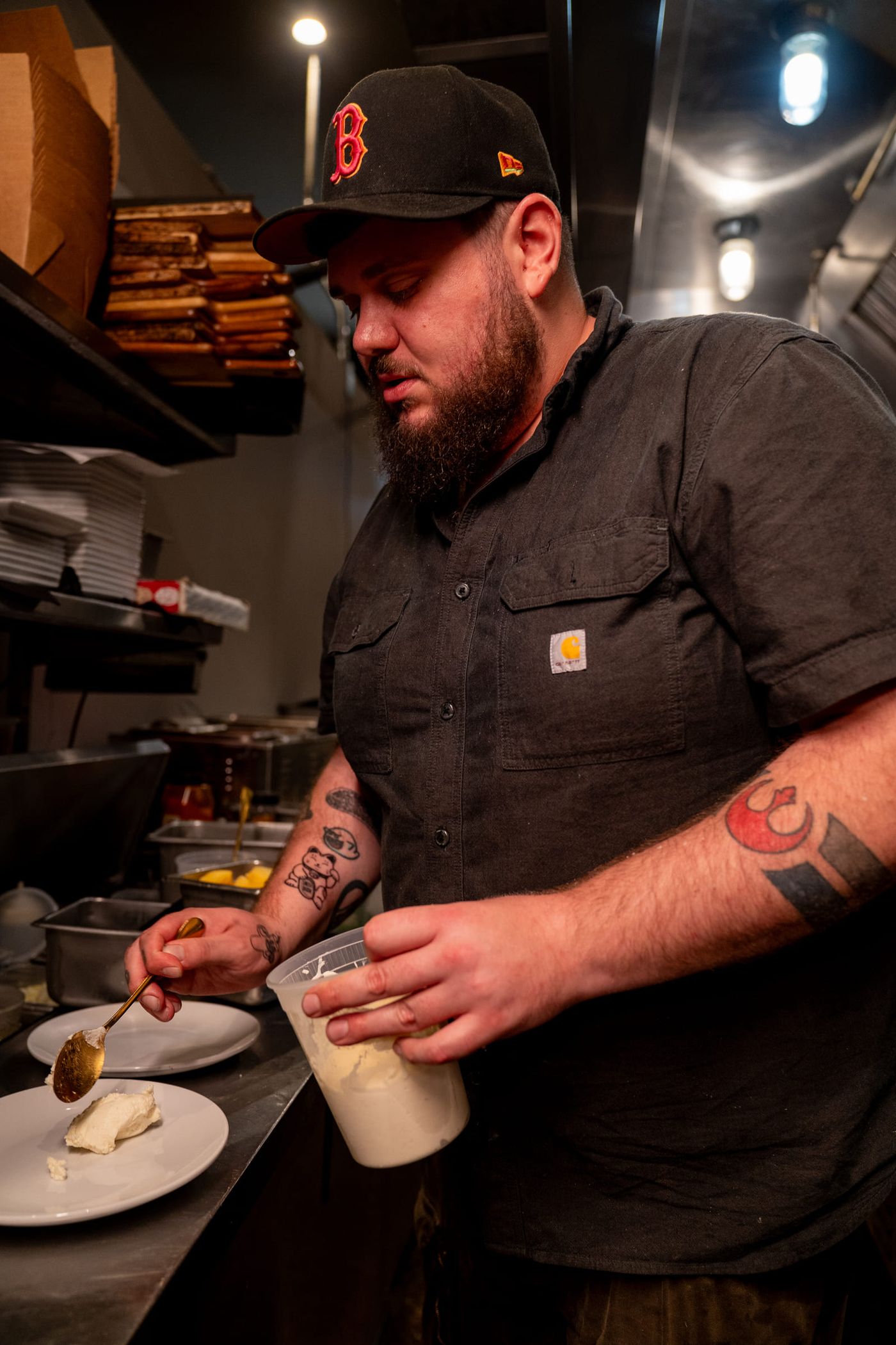 A bearded cook in a black cap plates a dessert with a spoon on a restaurant kitchen line.
