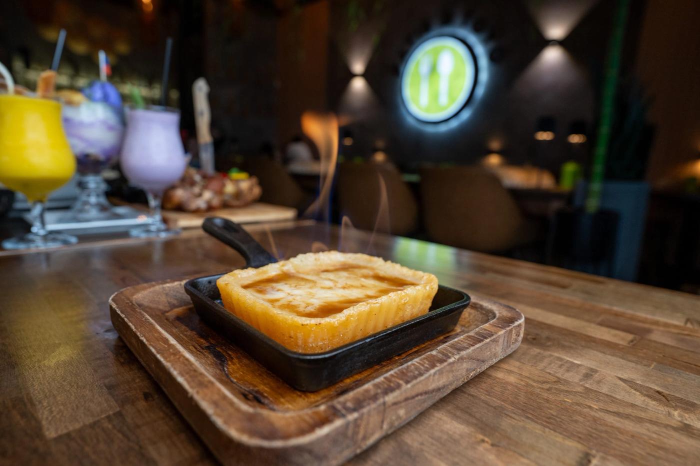 A golden baked dessert in a cast-iron skillet sits on a wooden board at a dim restaurant table, with colorful drinks and a glowing wall sign behind it.
