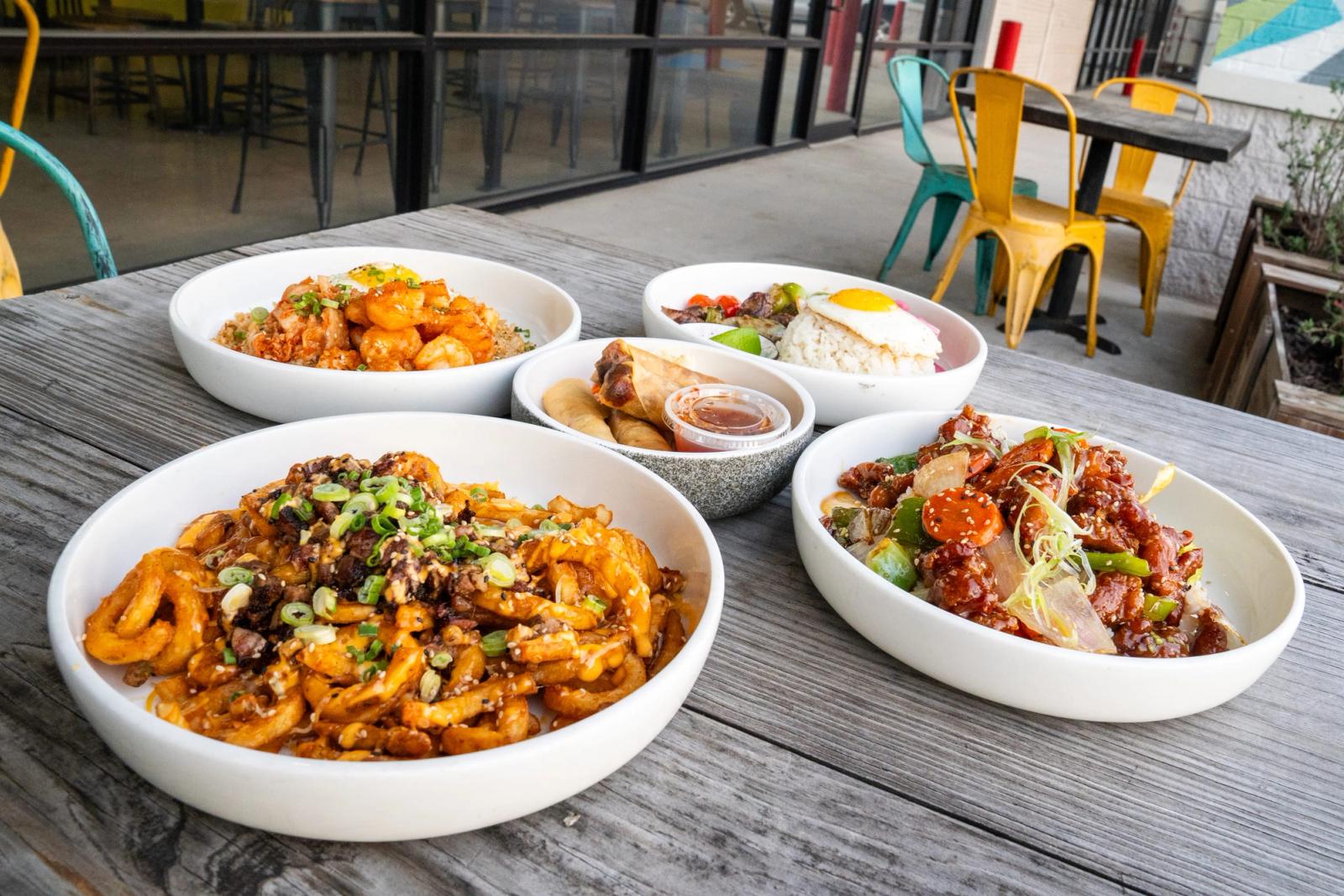 Five dishes, including loaded curly fries, egg rolls, rice bowls with fried egg and glazed chicken, set out on an outdoor patio table with colorful chairs.