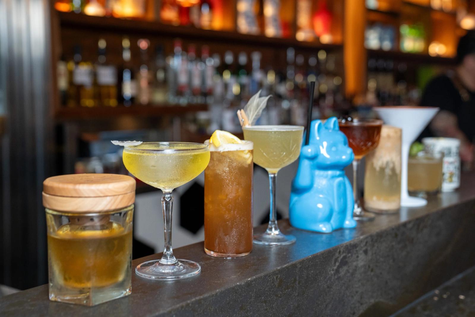 A row of assorted cocktails, including coupes, a highball, a lidded jar drink and a blue cat-shaped tiki mug, on a dark bar with bottle shelves behind.