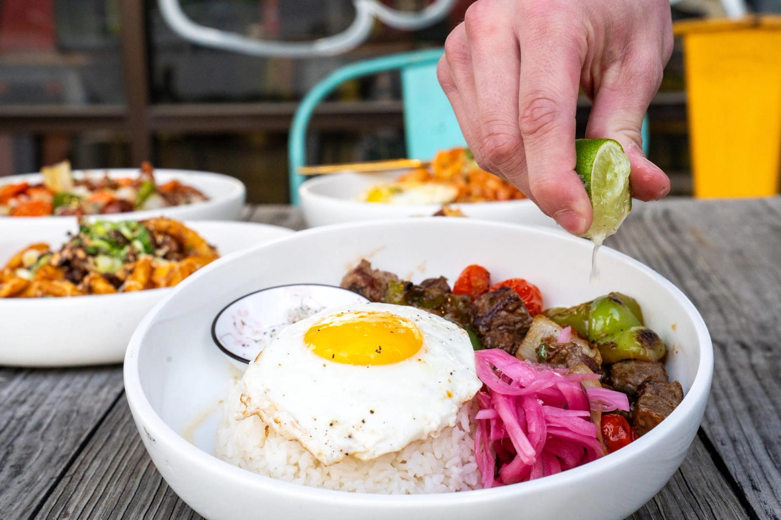 A hand squeezes lime over a rice bowl with a fried egg, pickled onions and beef on a patio table surrounded by other dishes.