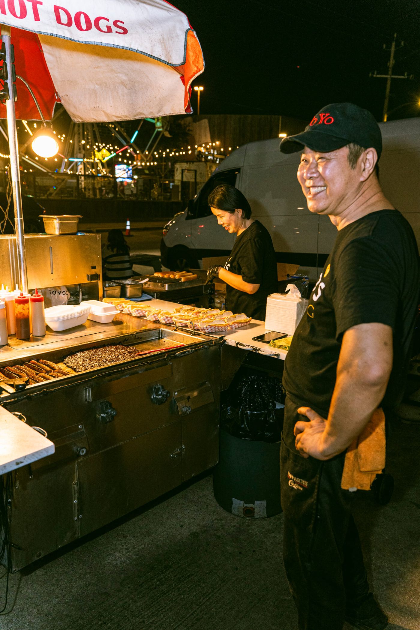 A smiling man in a black cap stands beside a hot dog cart at night while a woman works the grill behind him under string lights.