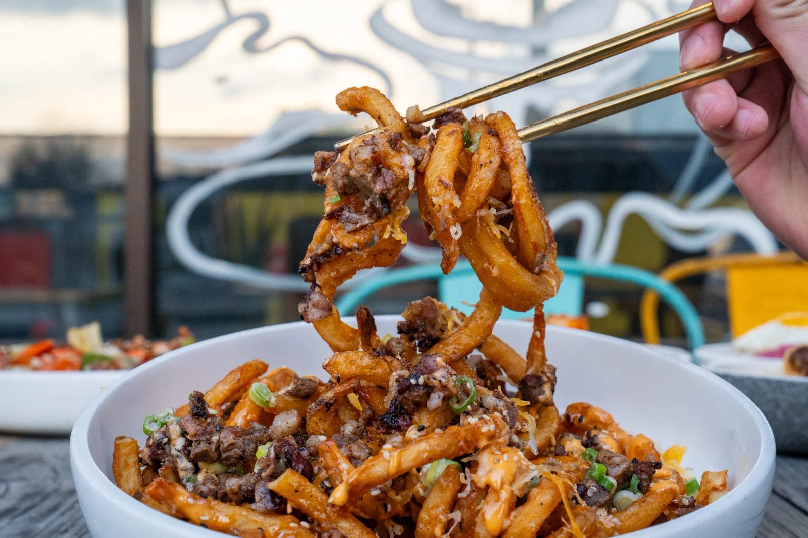 Chopsticks lift thick stir-fried noodles with ground meat and herbs from a white bowl on an outdoor patio with a mural behind.