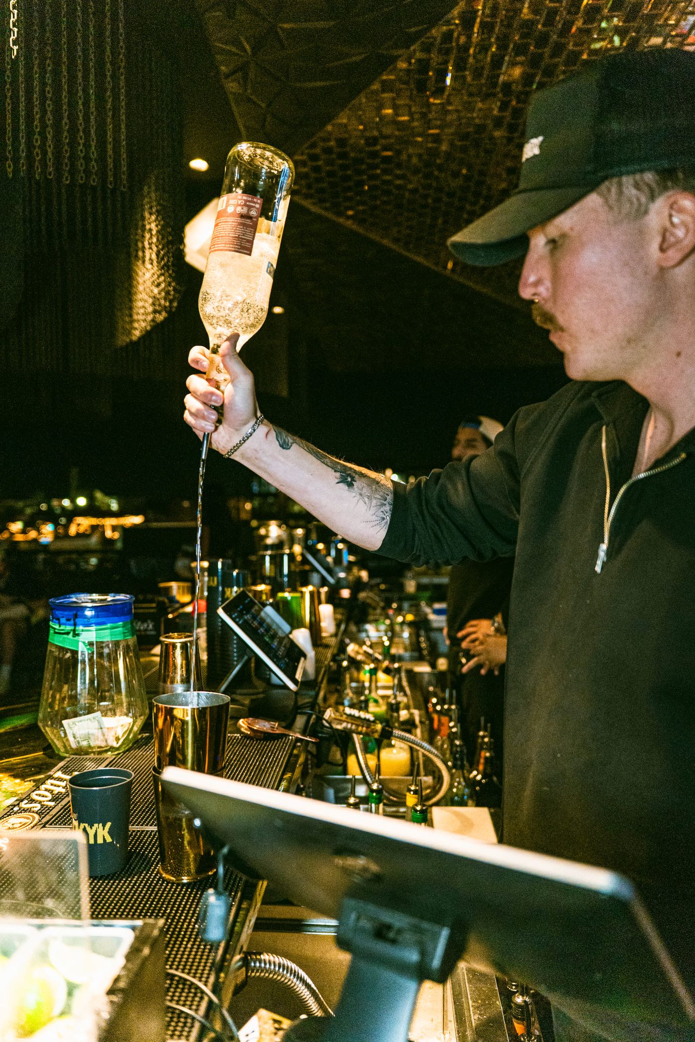 A bartender in a black cap free-pours from an upturned bottle into a shaker at a dark bar with a gold-tiled ceiling.