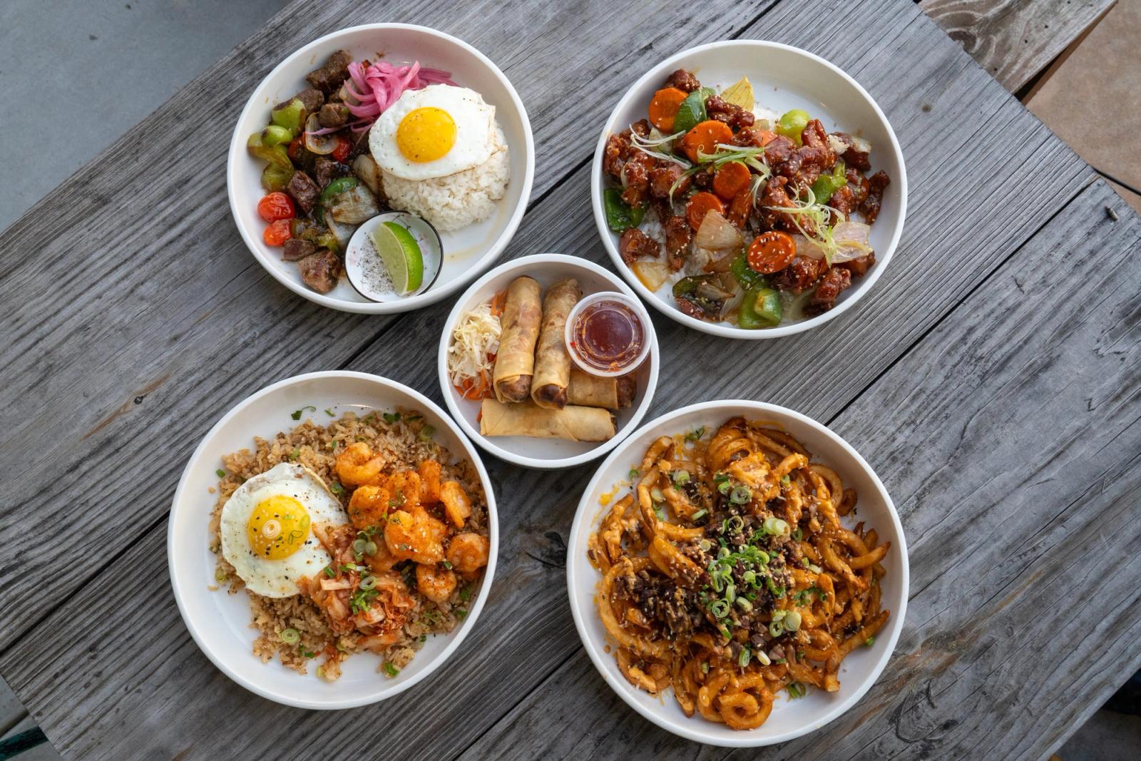 Five bowls on a wooden table, including a beef rice bowl with egg, shrimp fried rice, a beef and vegetable dish, spring rolls with dipping sauce and stir-fried noodles.